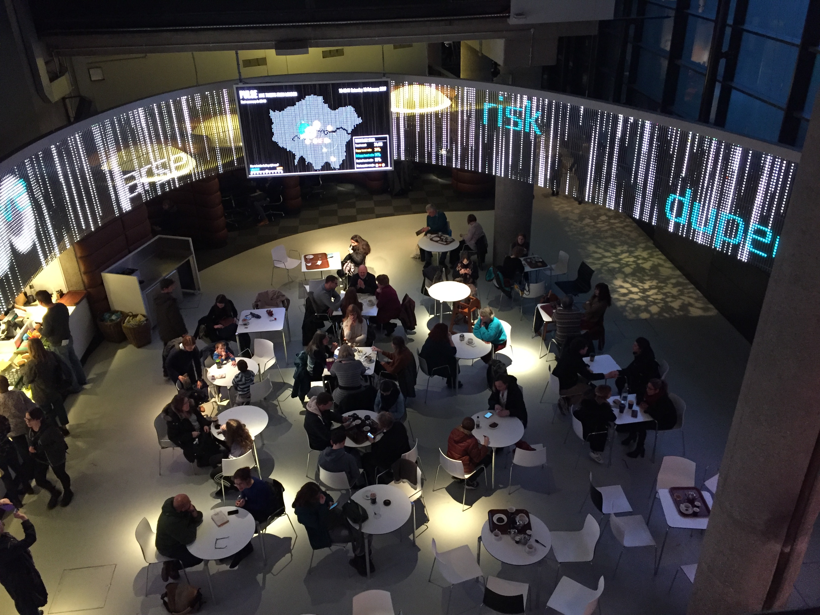 Museum café, with a computer screen stretching around the curved ceiling above. A large monitor in the centre shows a map of London. The screen stretching around the wall has the words risk, dupe and arse in random positions, over a variety of vertical lines.