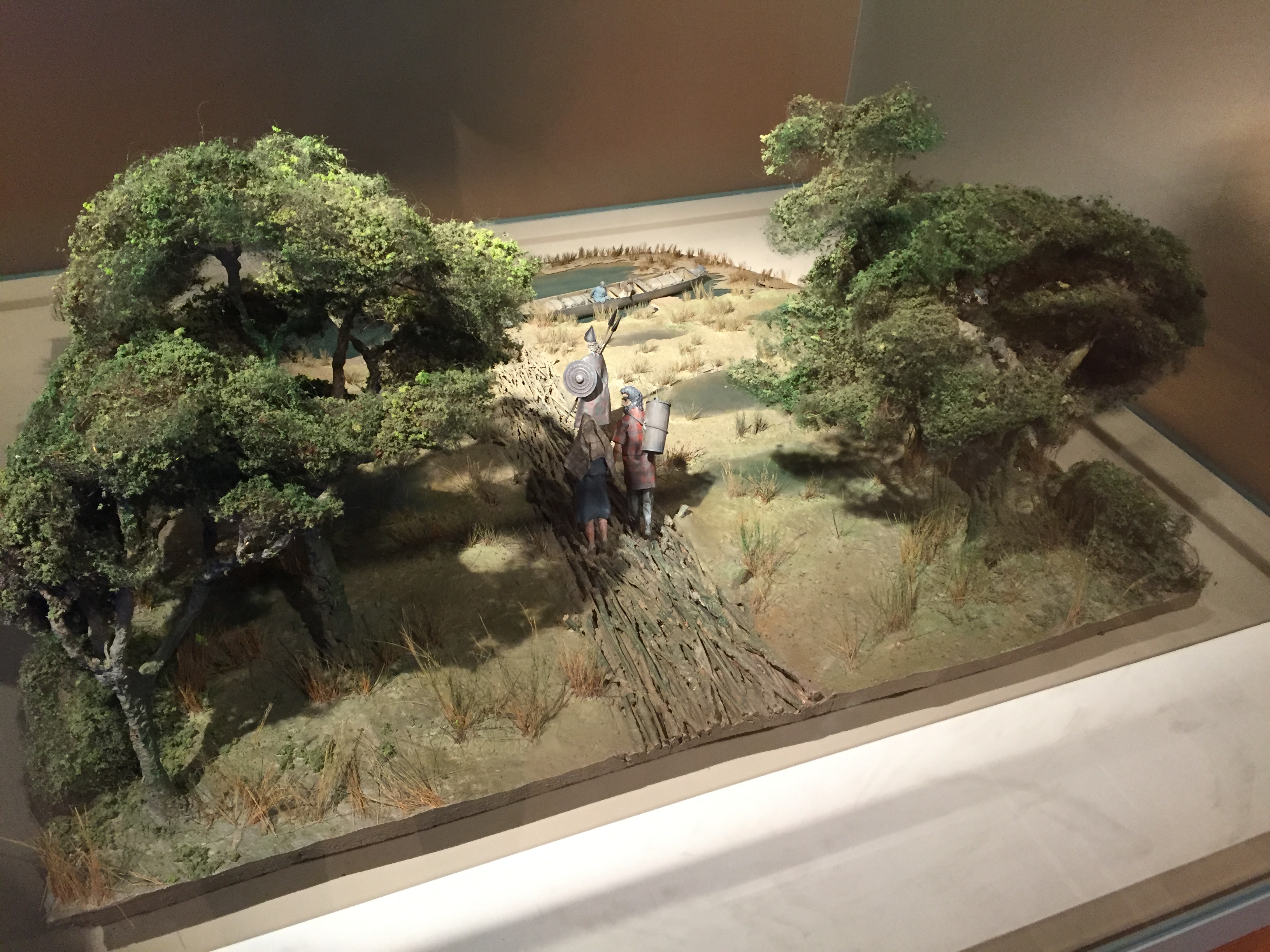 Model of a green landscape. A path between two sets of trees has models of people carrying camping gear towards a river or lake, where a long wooden boat is moored.
