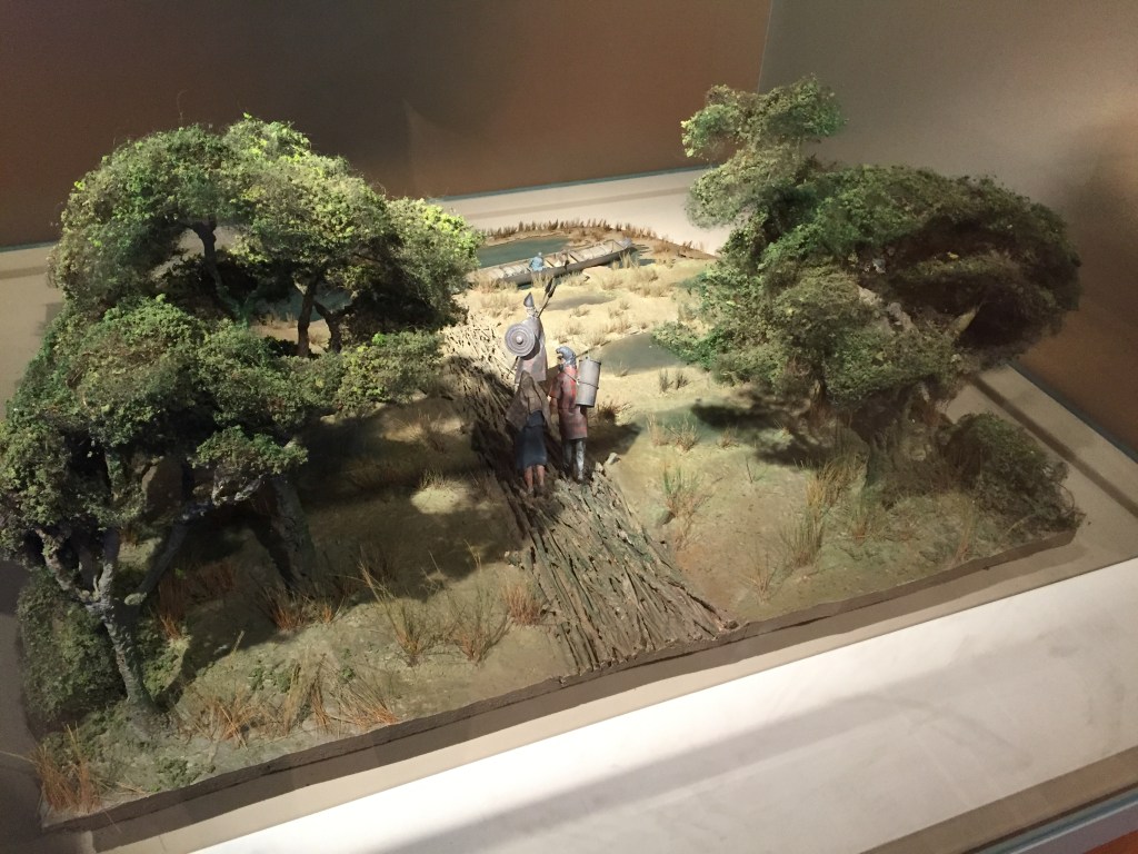 Model of a green landscape. A path between two sets of trees has models of people carrying camping gear towards a river or lake, where a long wooden boat is moored.
