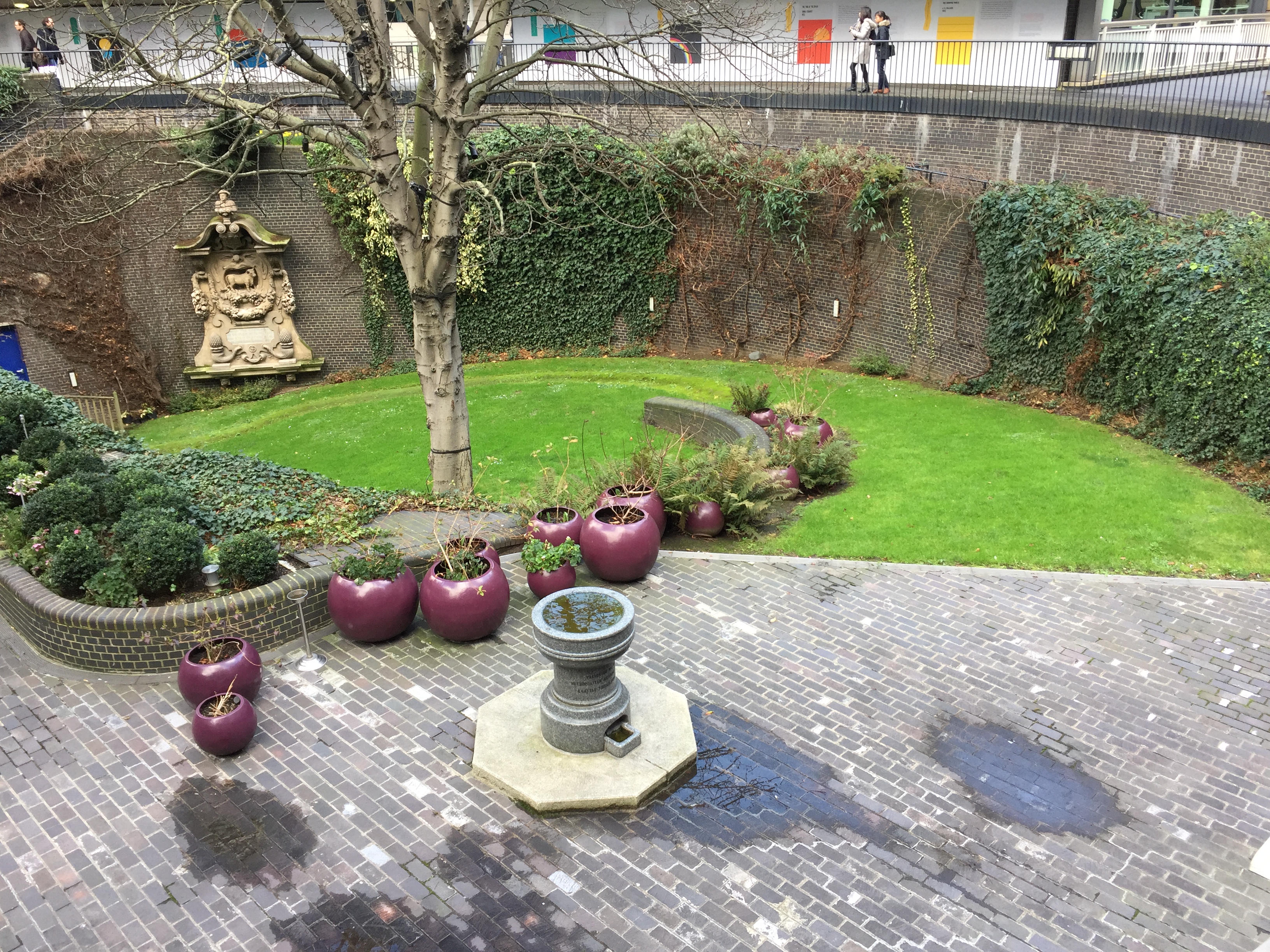 Large round garden area, half covered in grass and the other half with paving, with round red pots containing plants cross the dividing line. The garden is surrounded by a tall wall with greenery growing all over it, apart from one section that has a large gold sculpture on it.
