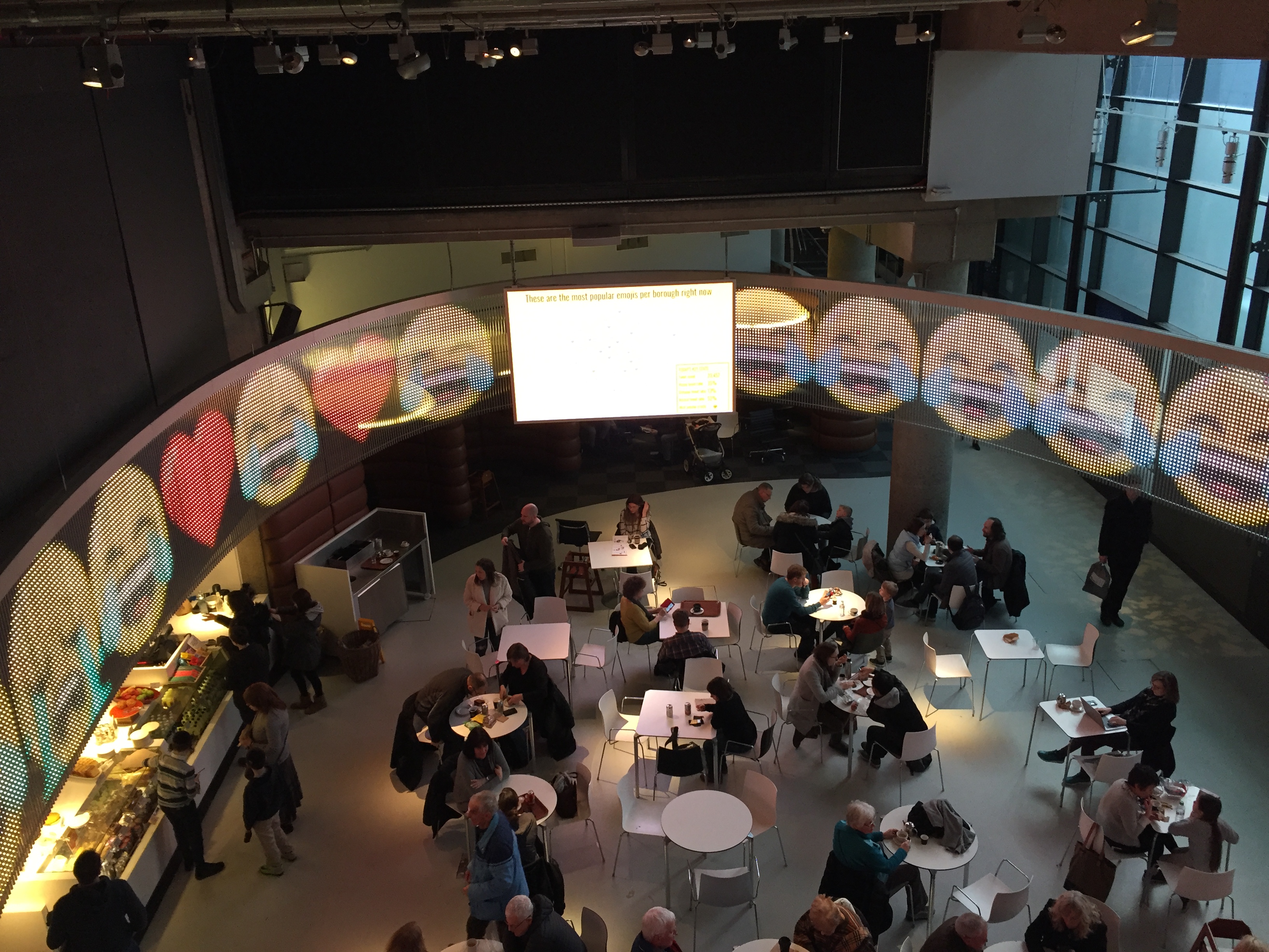 Museum café, with a computer screen stretching around the curved ceiling above, filled with large crying with laughter emojis.