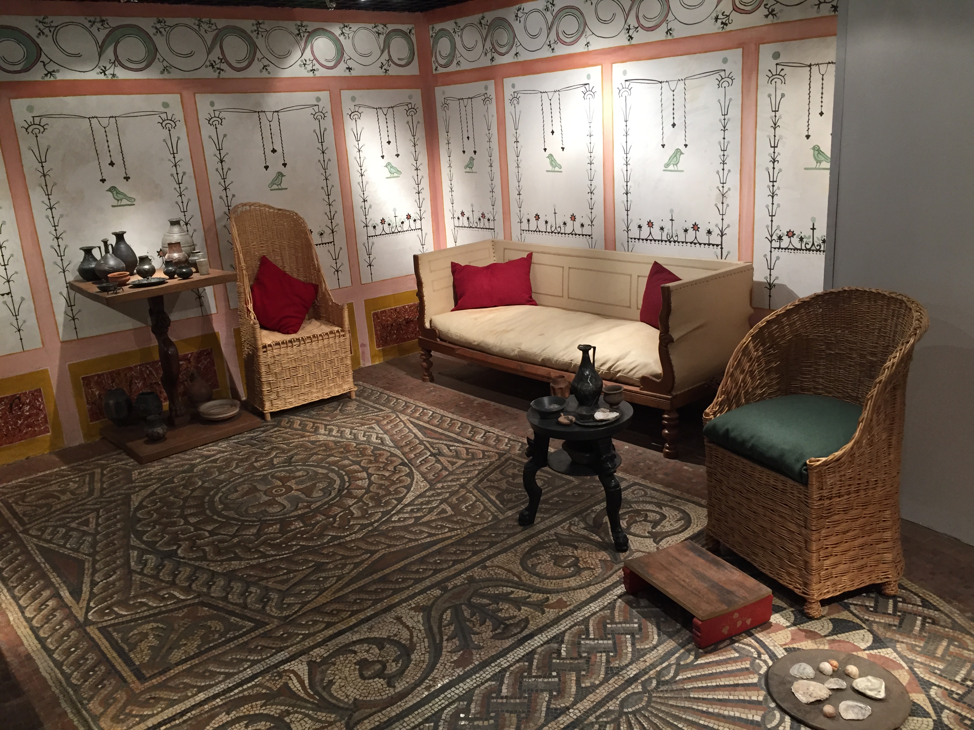 A furnished room containing a white 3 seater sofa, 2 chairs made of a wicker basket type material with tall, rounded backs, and an ornately decorated carpet.