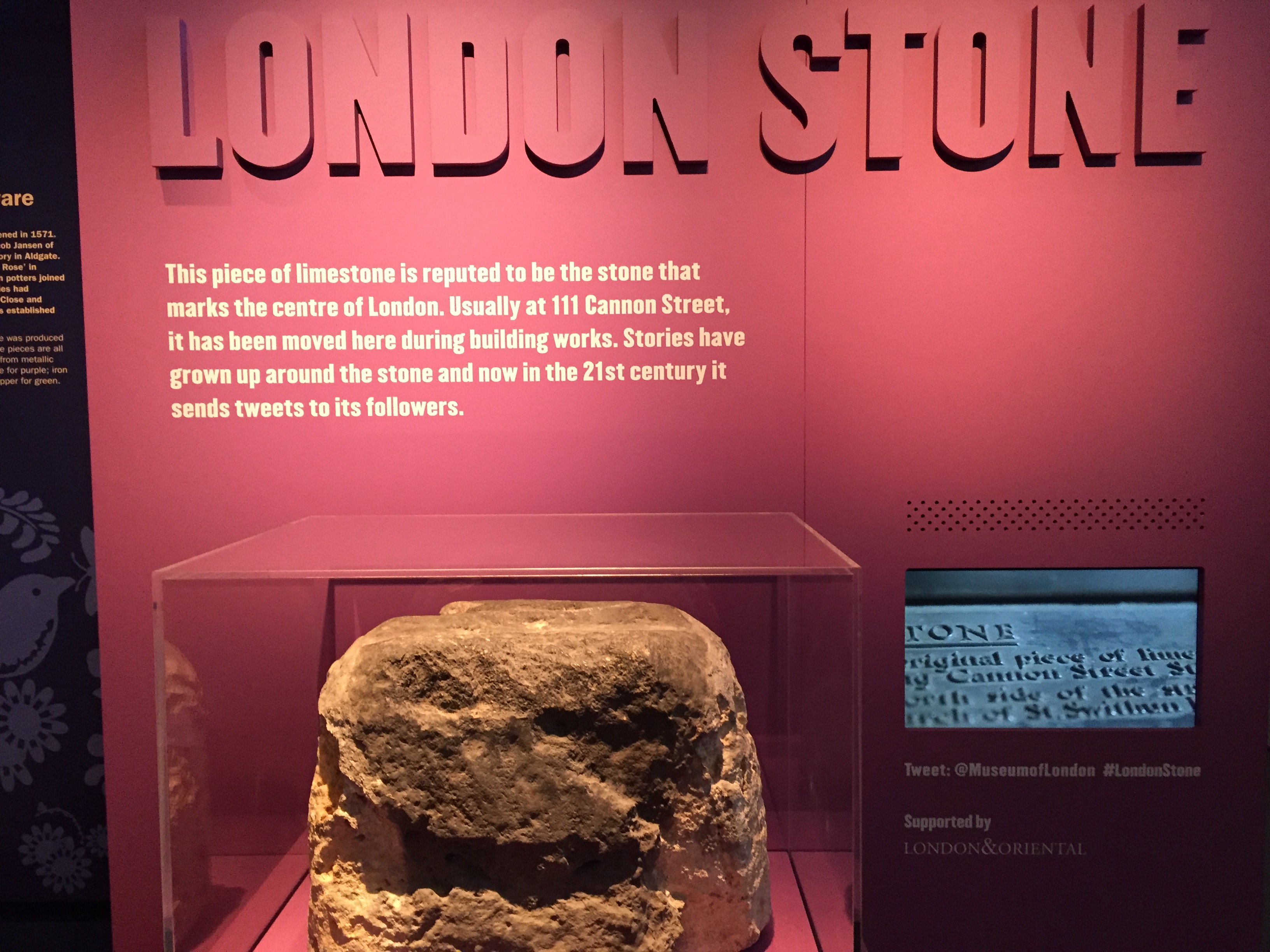 A large piece of limestone in front of a large dark pink panel with large bold white text, describing it as the London Stone that is reputed to mark the centre of London at 111 Cannon Street.