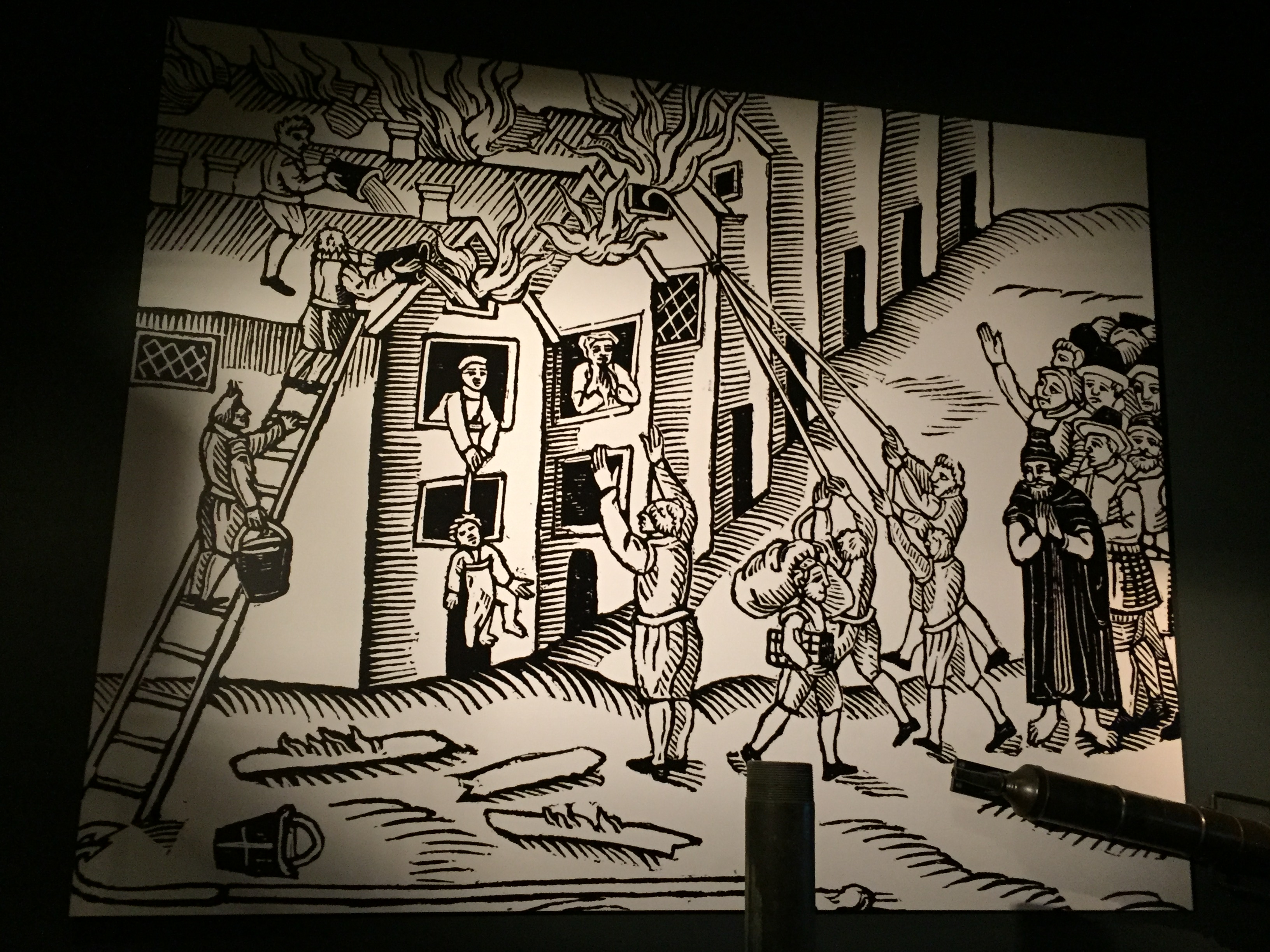 Black and white cartoon depicting the Great Fire Of London. A crowd watches as people with buckets of water climb a ladder to fight the fire on the roof of a house, while a person inside the house has a rope tied around their child's chest, and is attempting to lower them to safety through the window.
