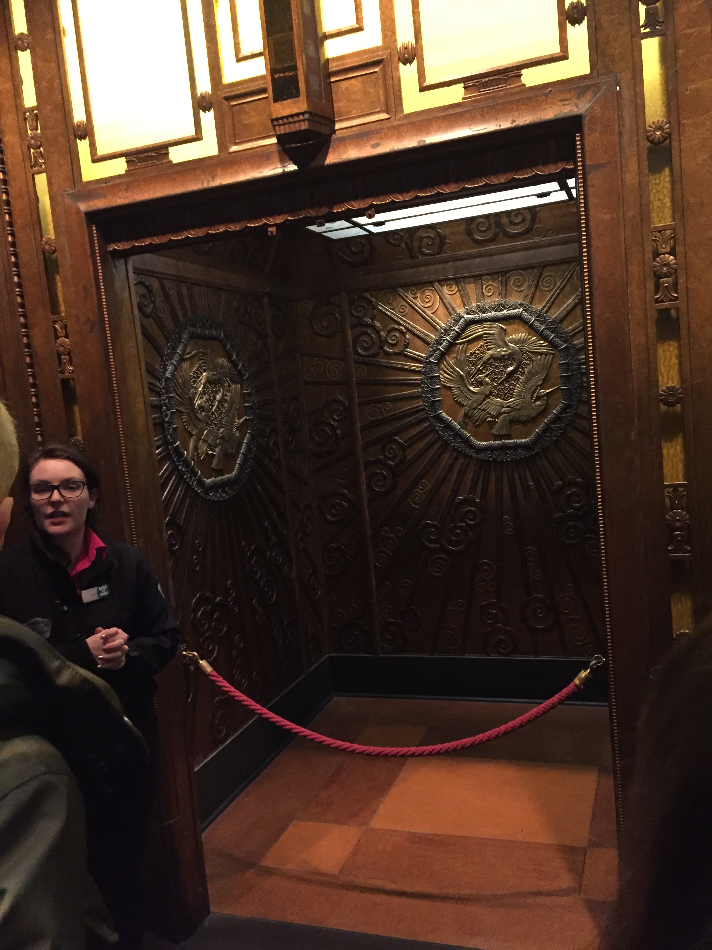 A female member of museum staff giving a talk in front of a big, open lift from Selfridges. The walls of the lift are ornately decorated, the notable feature being a large octagon shape containing 3 golden birds flying in a circular formation.