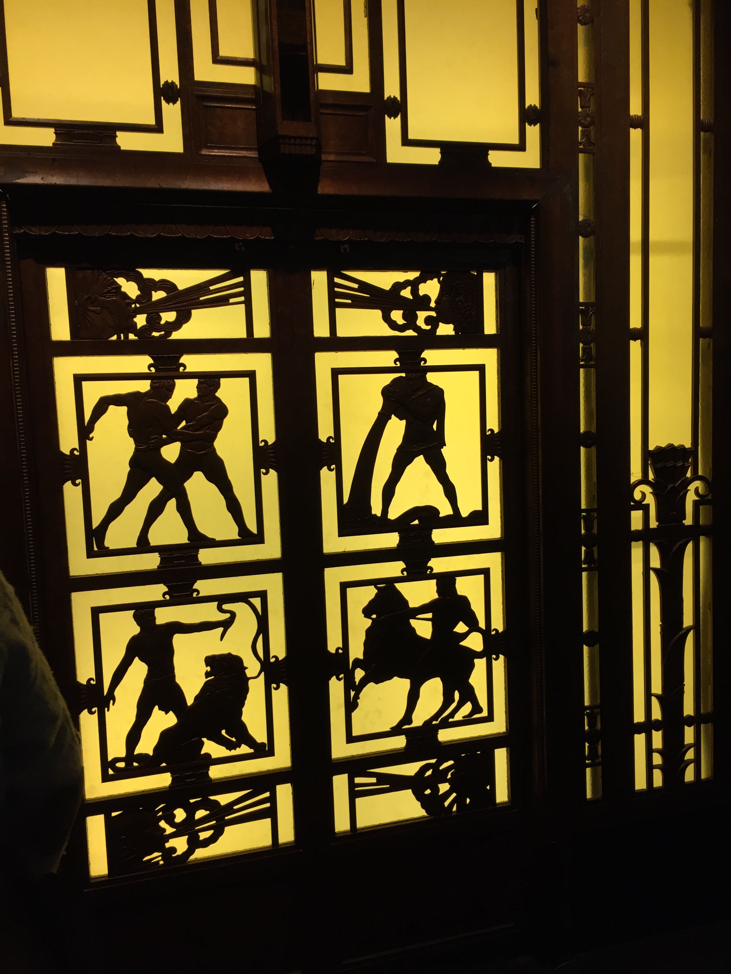 Doors of the Selfridges lifts. The doors have black squares against a yellow background, and inside the squares are black silhouette of people doing things like fighting, taming a lion and riding horseback.