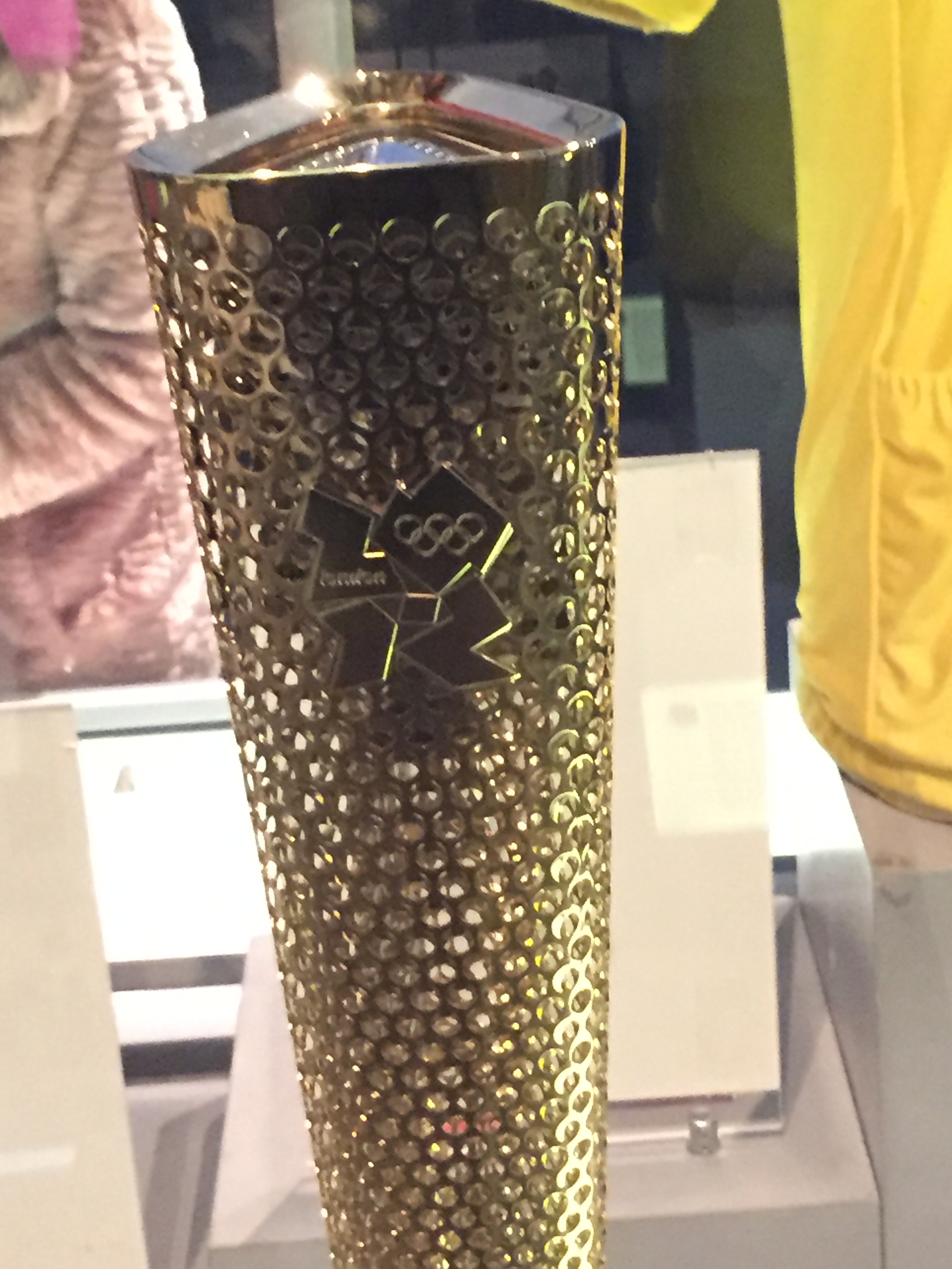 London 2012 Olympic Torch, tall and gold with small round holes all over it, and the 2012 Olympic logo embossed on it.