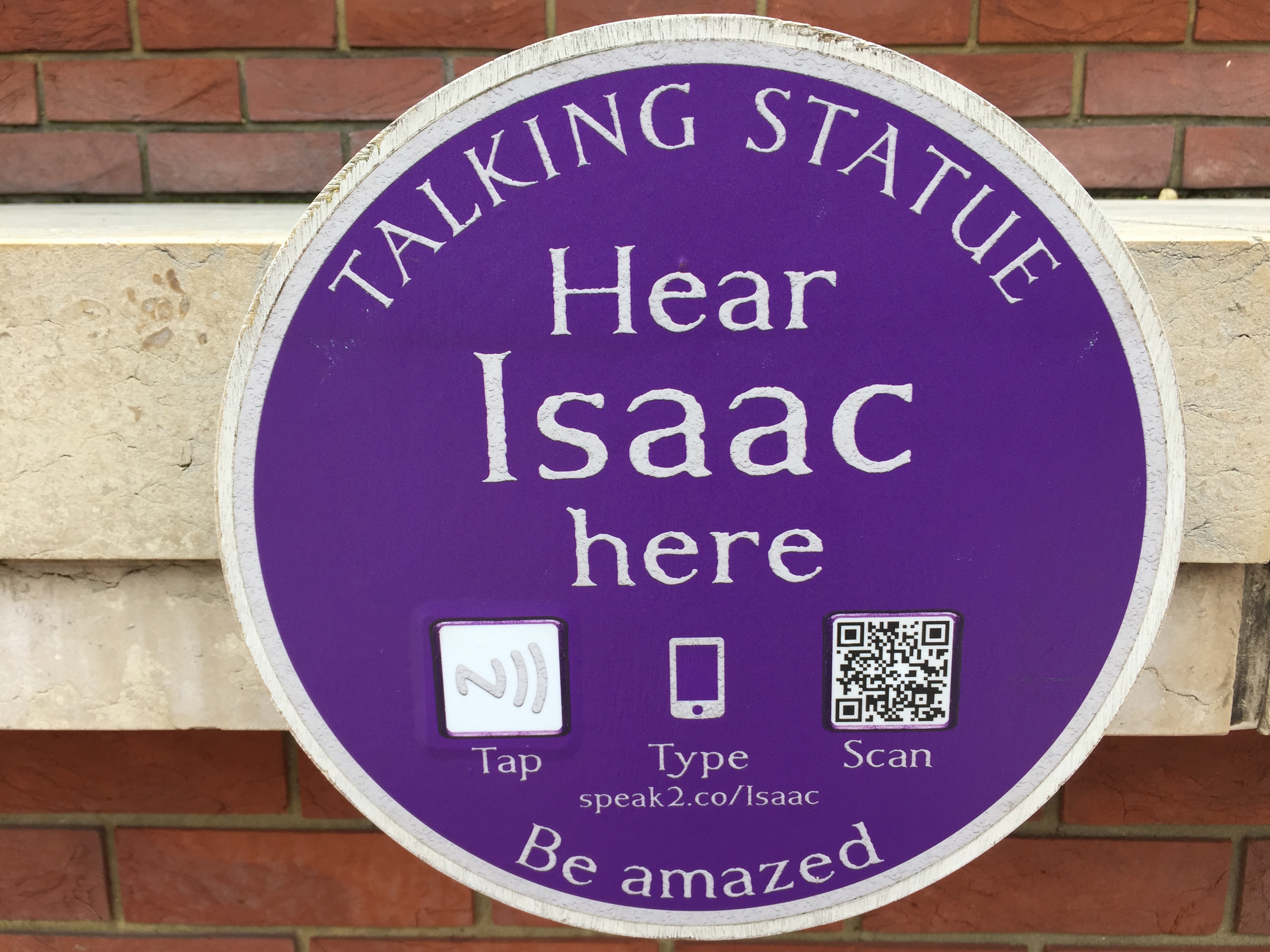 Circular blue plaque with white text that says Talking Statue, Hear Isaac here, Be Amazed. 3 icons invite the reader to either tap the plaque with a phone that can read the chip inside it, or scan a QR code, or manually type in a short web address - speak2.co/Isaac