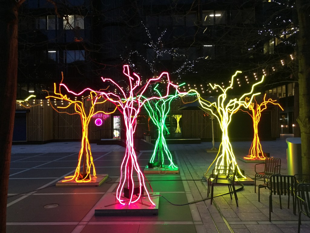 Five tree-like sculptures made up of wavy lights that spread out at the top like branches and out at the bottom like roots. Each tree is lit in a different colour - from left to right, they are orange, pink, green, yellow and orange again.
