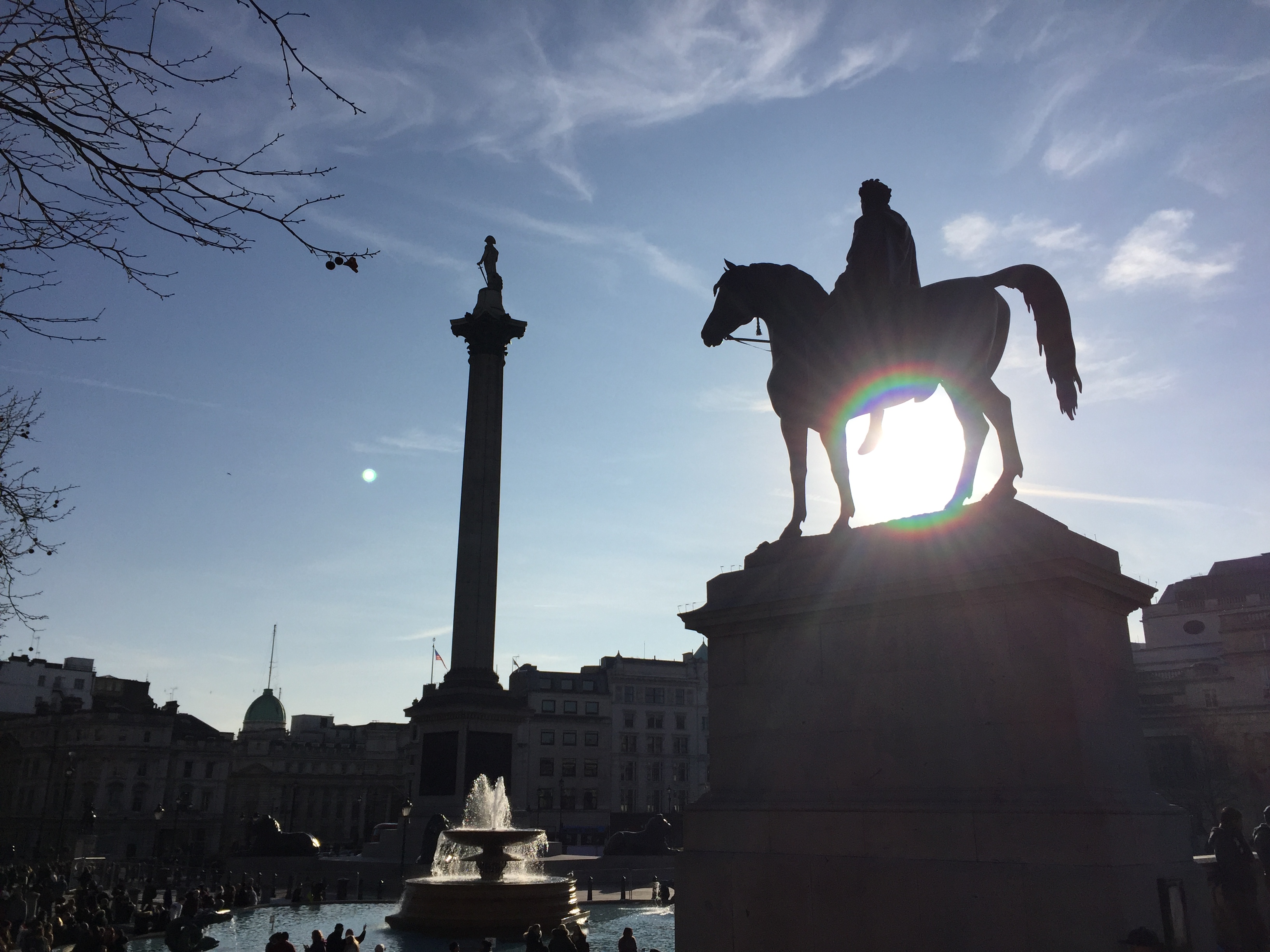 Trafalgar Square. Nearest to the camera, on the right, is a square plinth, on top of which is a statue of a man on a horse, with the sun shining through the space between the horse's front and back legs. To the left, further away, is Nelson's column towering over a circular fountain.
