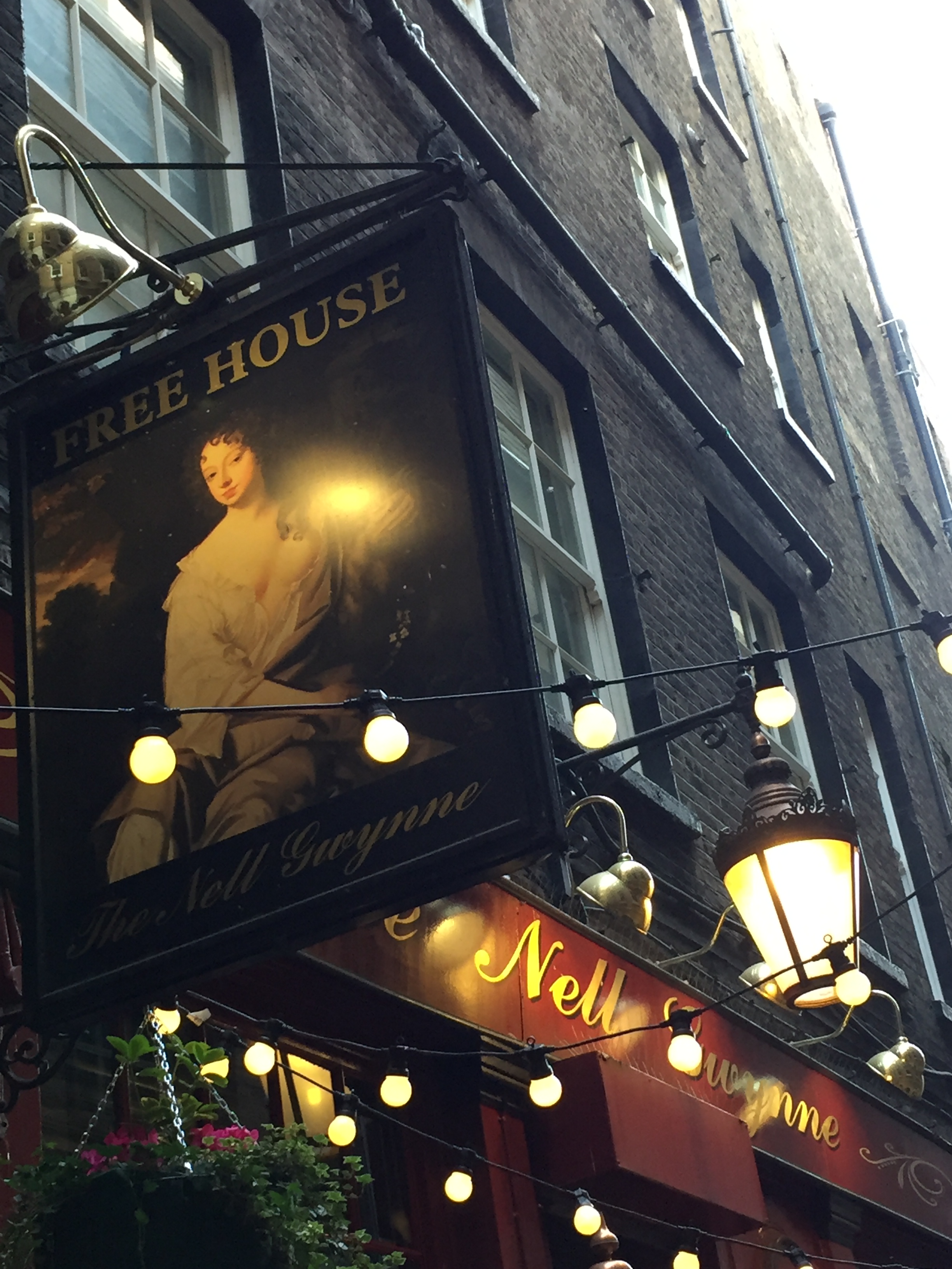 Exterior of Nell Gwynne's Tavern, including a sign featuring a portrait of Nell below the words Free House. Strings of lights also cross the front of the pub, and an old fashioned lantern style light hangs out from behind the portrait sign.