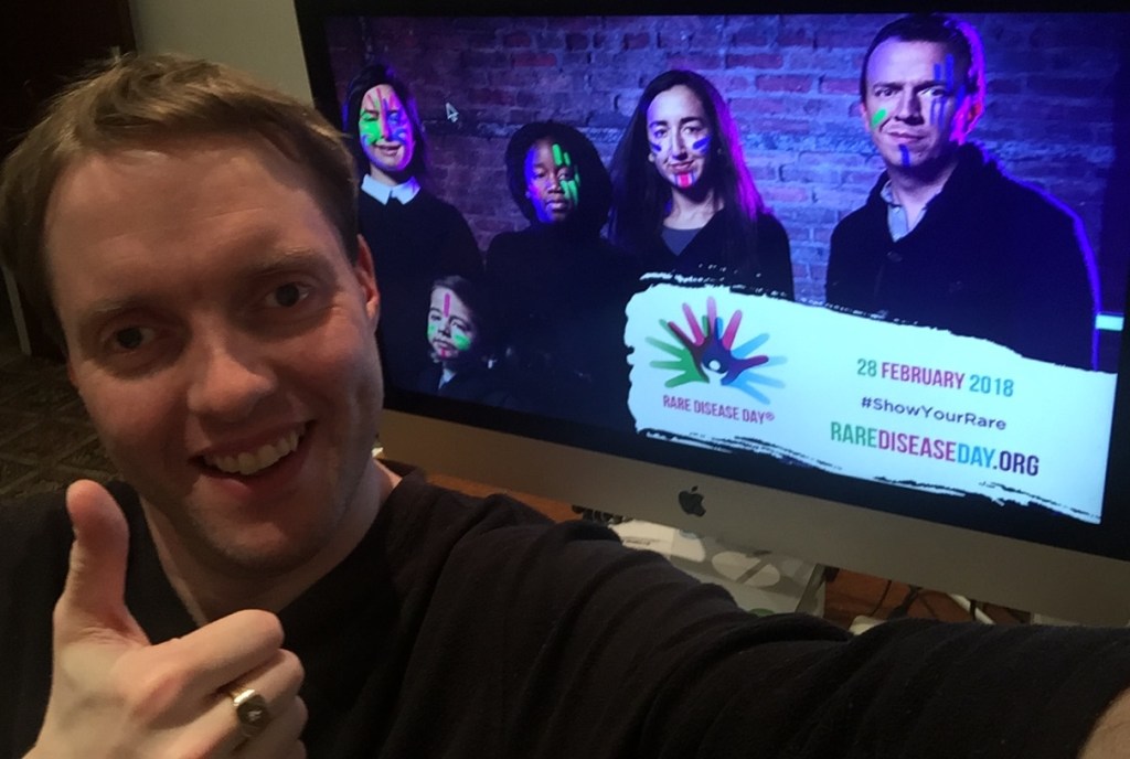 Me smiling and giving a thumbs up, in front of the Rare Disease Day promotional image on my computer. The promo image shows 5 people wearing luminous face paint, and a banner in the corner says Rare Disease Day, 28 February 2018, Show Your Rare, RareDiseaseDay.org.