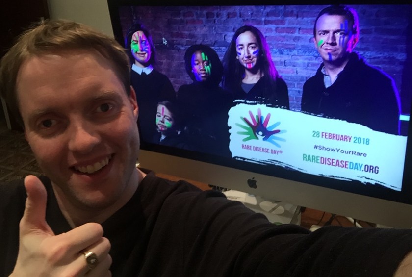 Glen smiling and giving a thumbs up, in front of the Rare Disease Day promotional image on his computer. The promo image shows 5 people wearing luminous face paint, and a banner in the corner says Rare Disease Day, 28 February 2018, Show Your Rare, RareDiseaseDay.org.