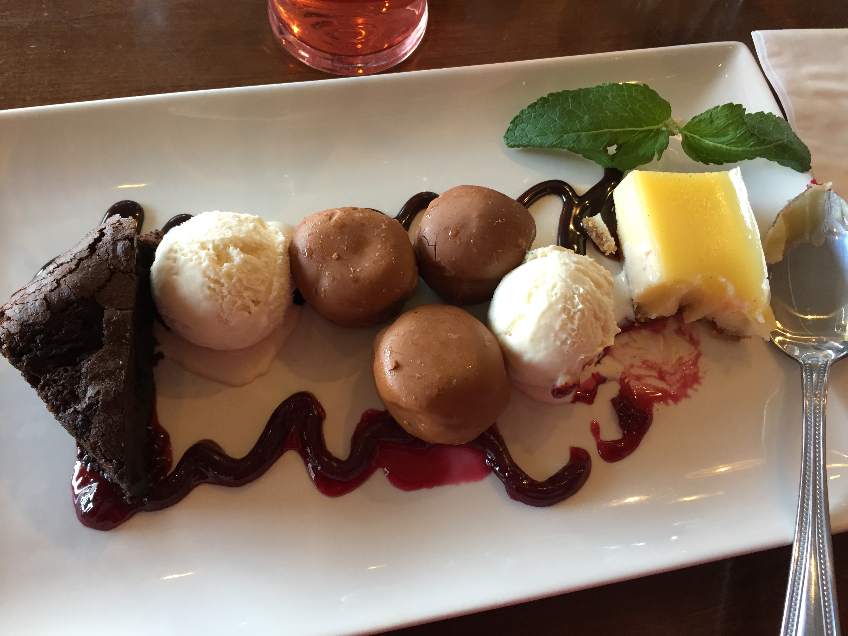 Rectangular plate with a slice of chocolate brownie, a scoop of vanilla ice cream, 3 chocolate profiteroles, another scoop of vanilla ice cream, and a slice of lemon tart, surrounded by swirling lines of strawberry sauce.