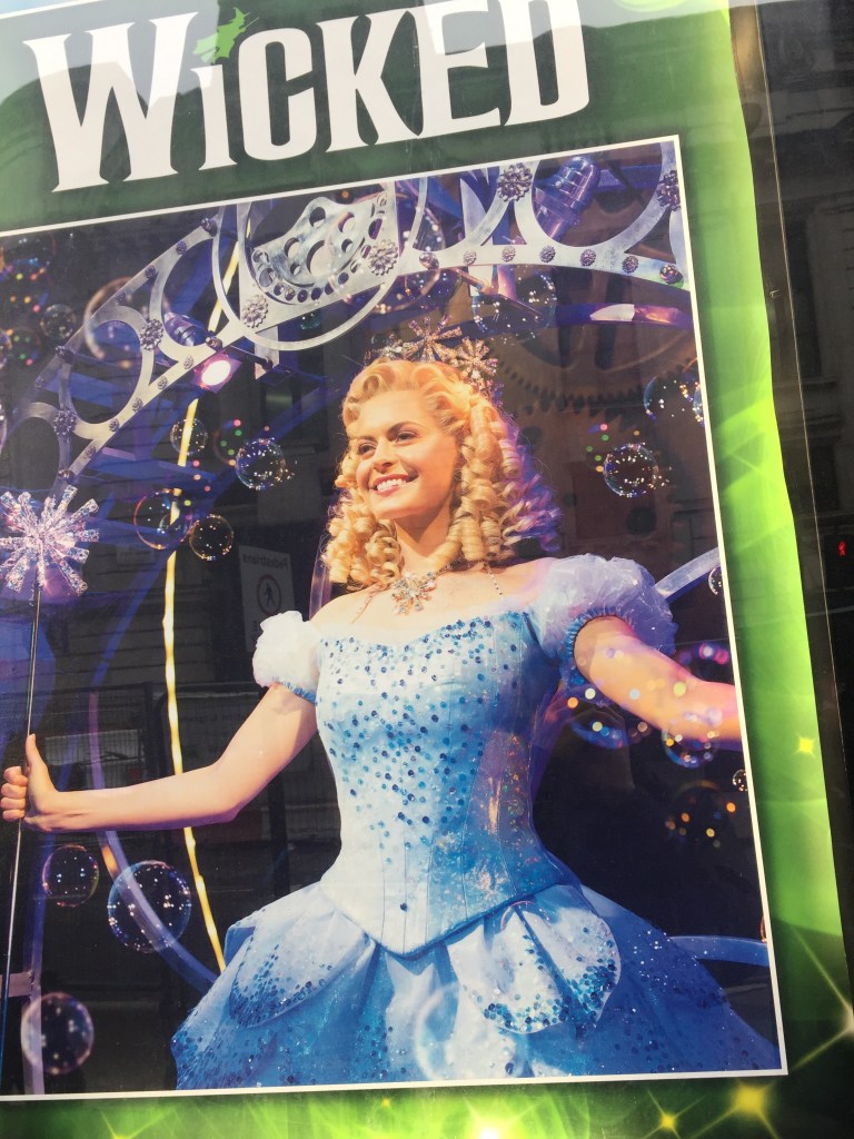 Poster showing the blonde, smiling Good Witch Glinda in her big blue dress, covered in sequins, and holding a wand with a large spiky silvery head on it.