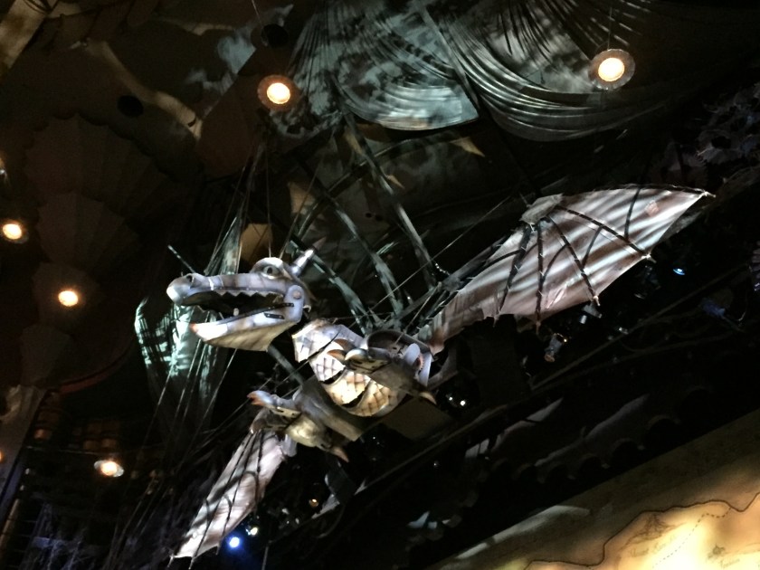 Large silver dragon above the stage, its wings spanning the entire width of the space, and its large head with open mouth looking straight ahead over the audience.