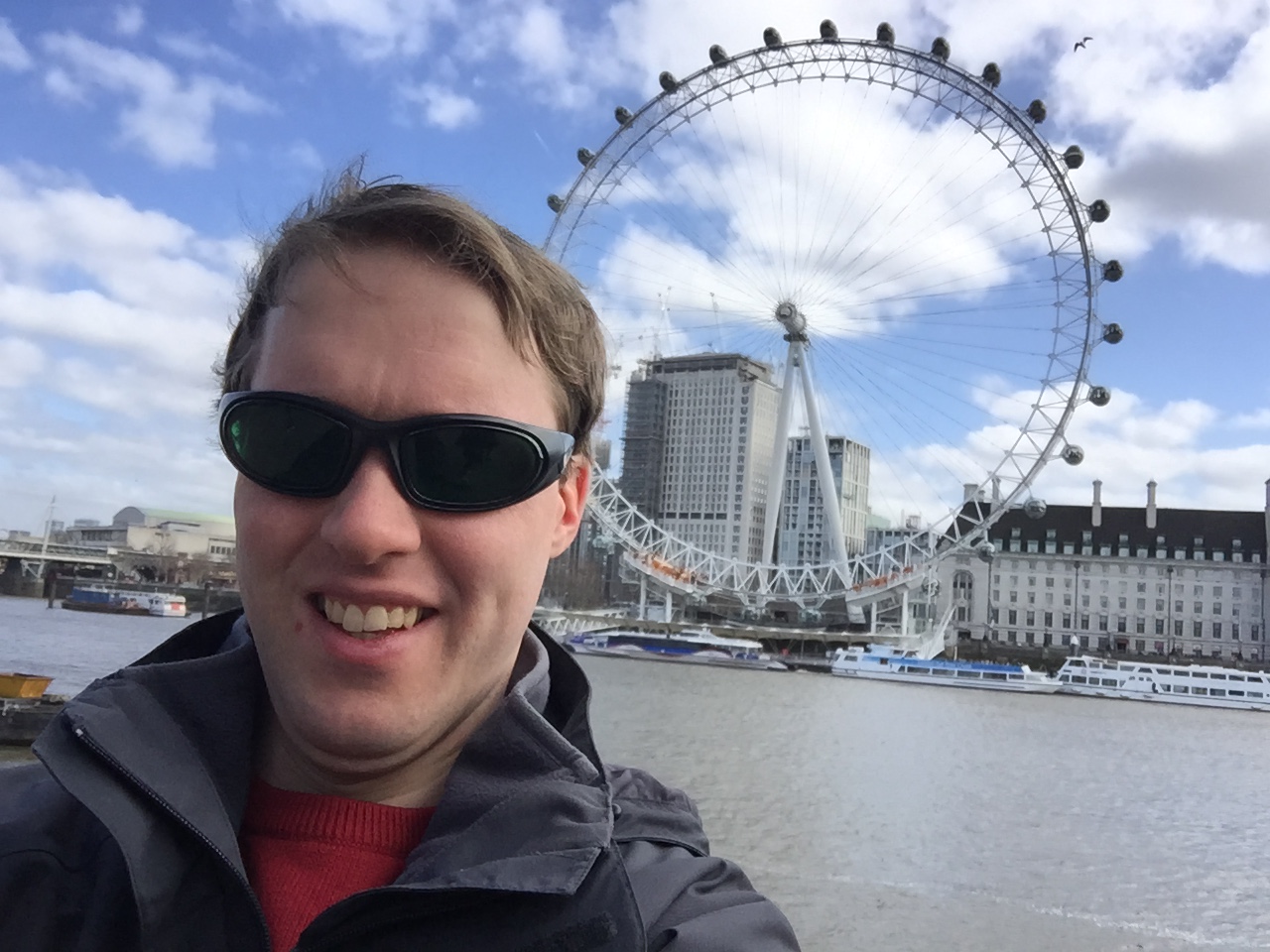 Selfie of Glen smiling and wearing sunglasses, with the London Eye behind him on the opposite side of the River Thames.
