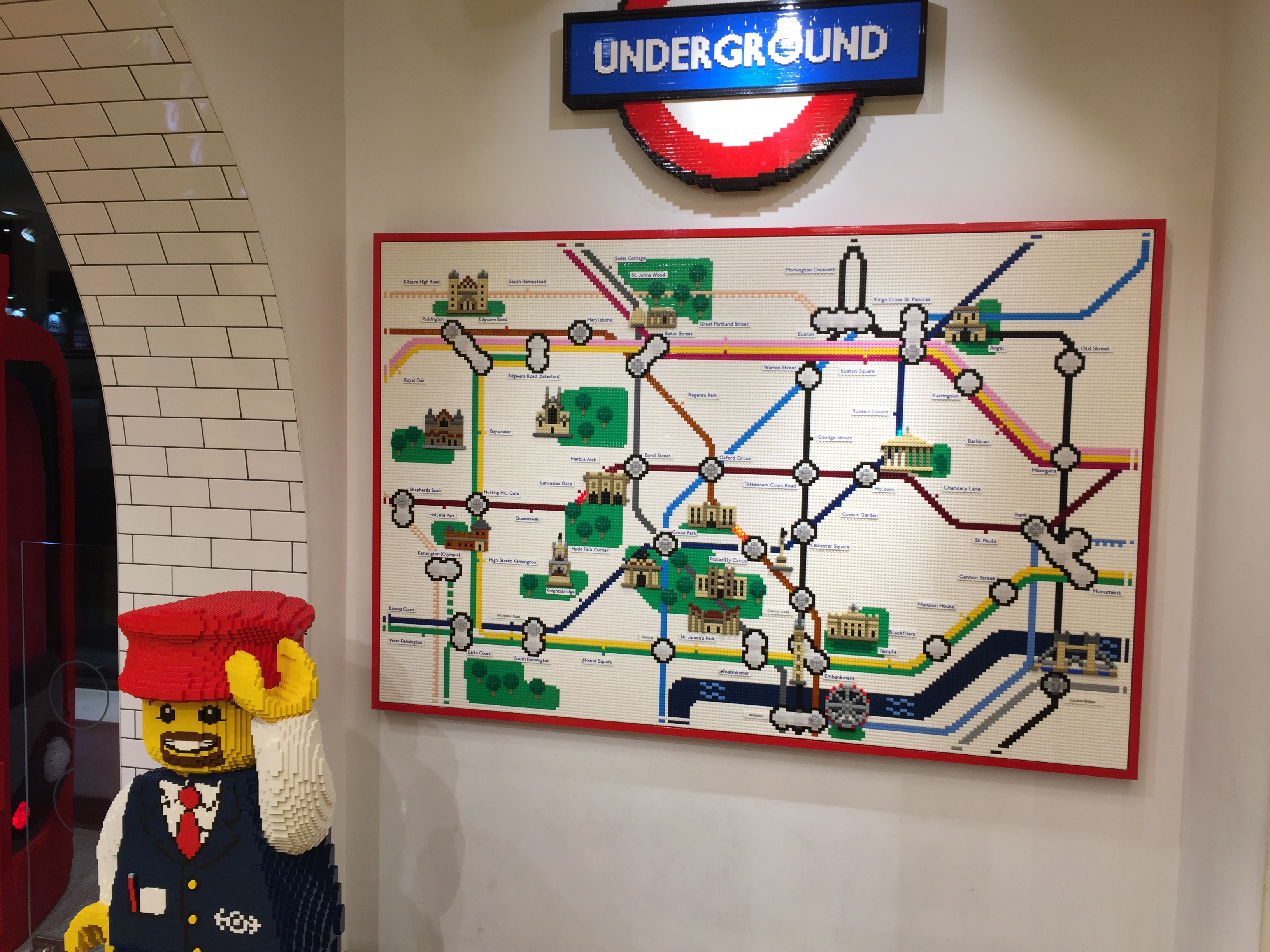 Lego version of the central section of the London Underground Map, with a Lego Underground roundel sign above it, and a Lego model of a smiling train guard on the floor in front of it.