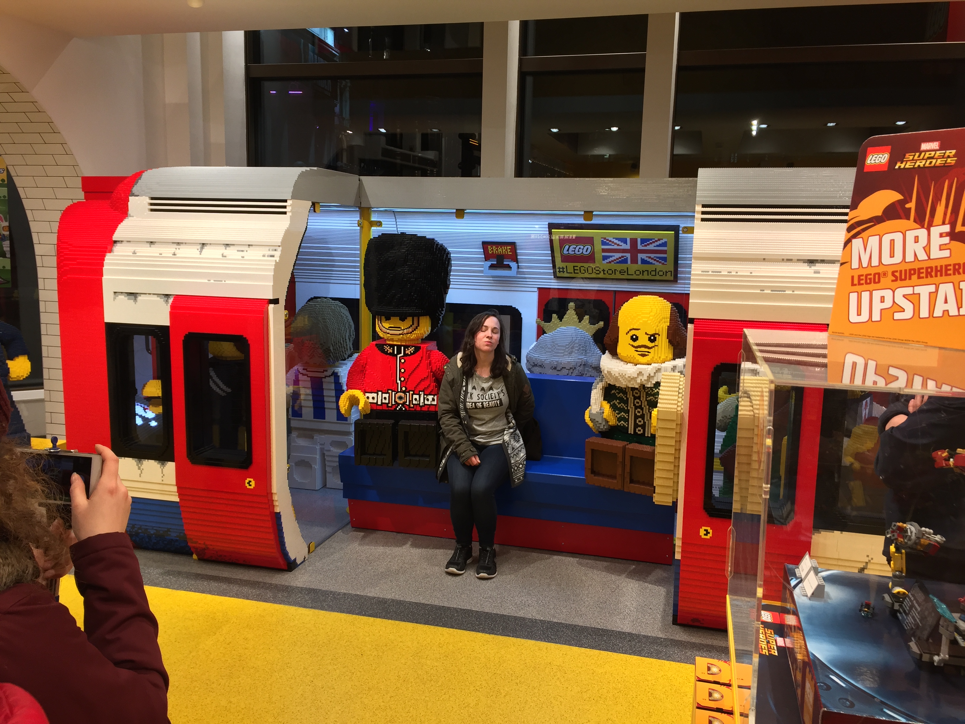 Large Lego model of a London Underground train carriage with its doors open. A Lego guard and a Lego Shakespeare are sitting inside, and a real-life woman is sitting in between them to have her photo taken.