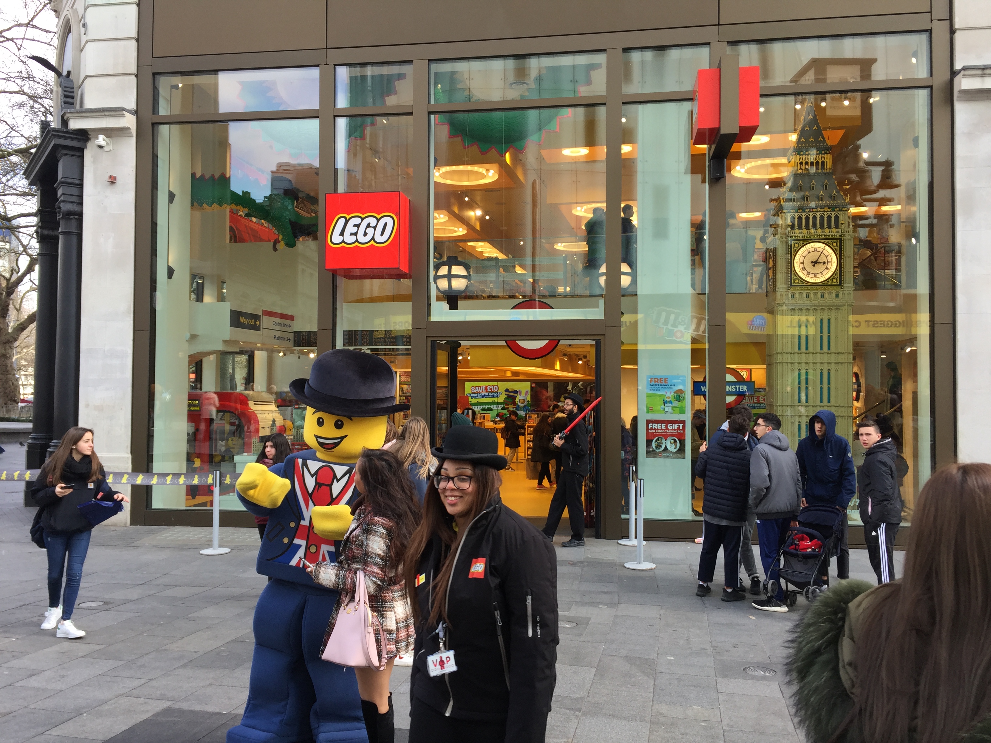 Outside the Lego Store, someone dressed as a lifesize yellow Lego Man, in a blue suit, Union Jack shirt and black bowler hat, poses with a lady member of the public for a photo. Next to them is a smiling female member of staff, wearing a black jacket with the Lego logo and black bowler hat.