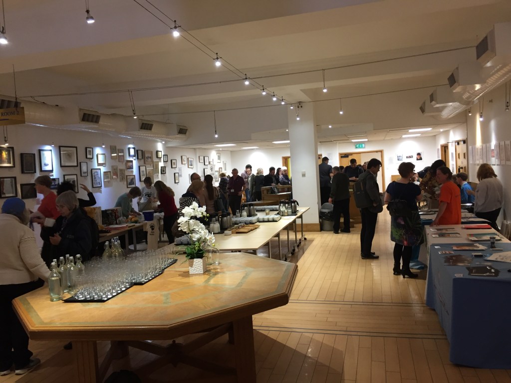 People gathering in the meeting area at the conference, with exhibition tables around the edge and drinks on the large central tables.