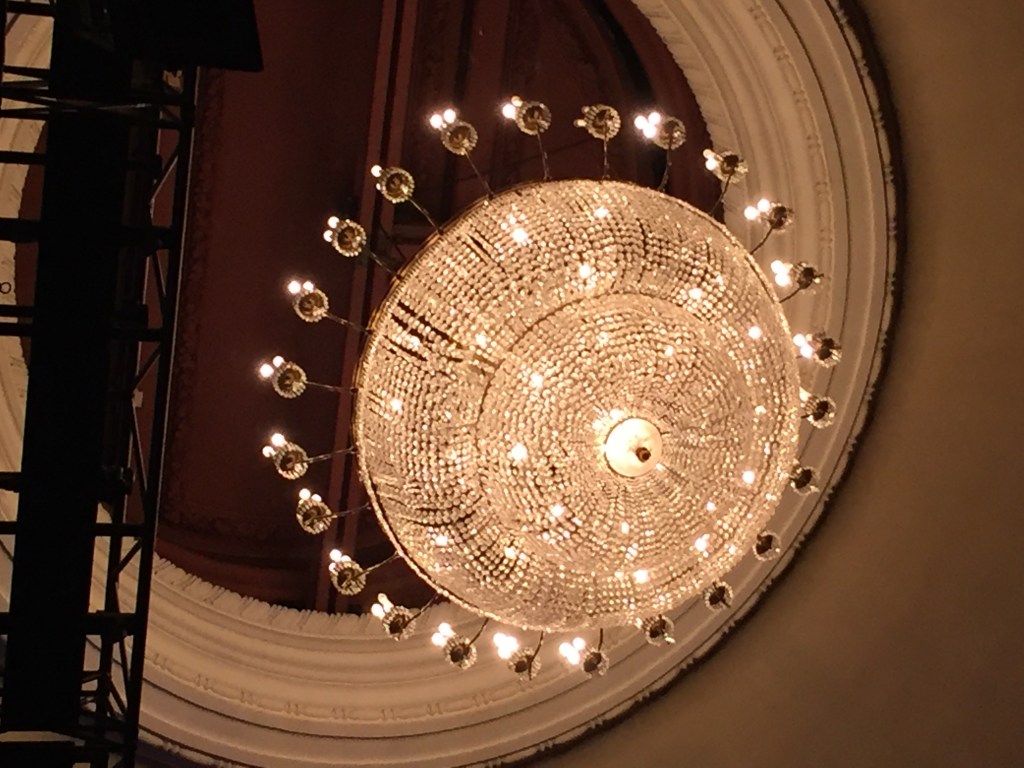 Ornate round chandelier light in the theatre auditorium, with lots of little lights sticking out all around it.