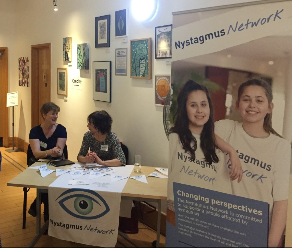 Sue Ricketts talking to a lady at the Nystagmus Network table ,with a tall poster next to it giving details of the charity.