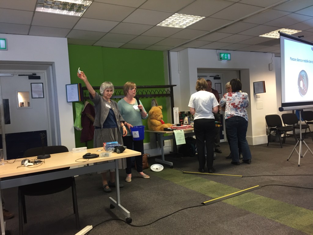 Raffle draw at the conference. One lady holds up the ticket drawn from the bucket held by the lady next to her. Among the prizes on the table next to them is a large cuddly teddy bear.