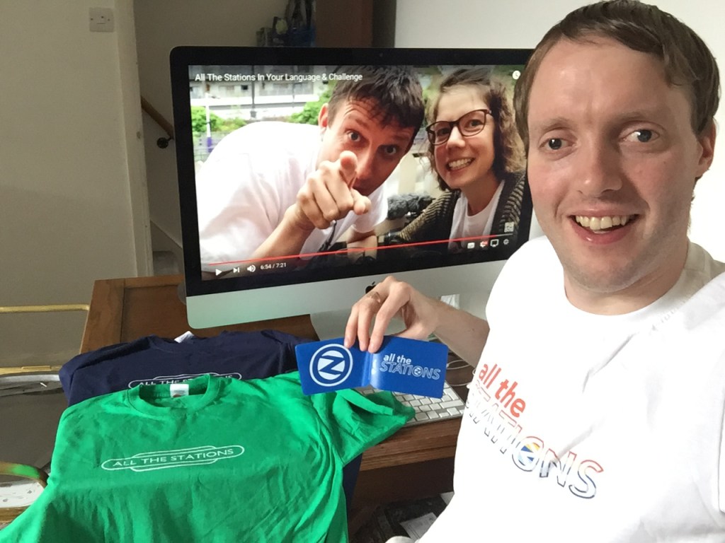 Me sitting at my computer, wearing a white All The Stations t-shirt, and holding a blue All The Stations Oyster card wallet. On the table next to me are 2 All The Stations t-shirts, one green and one navy. And on the computer screen, the Have An Adventure video is paused with Geoff and Vicki looking and pointing at the viewer through the camera.