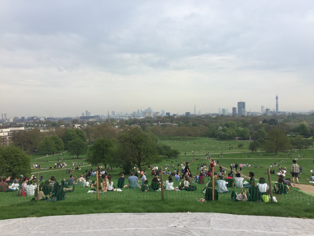 London skyline viewed from Primrose Hill. Lots of people are sitting on the grassy hill stretching down into the park below, while in the distance is the London skyline, including iconic sights like the Shard, BT Tower and London Eye.