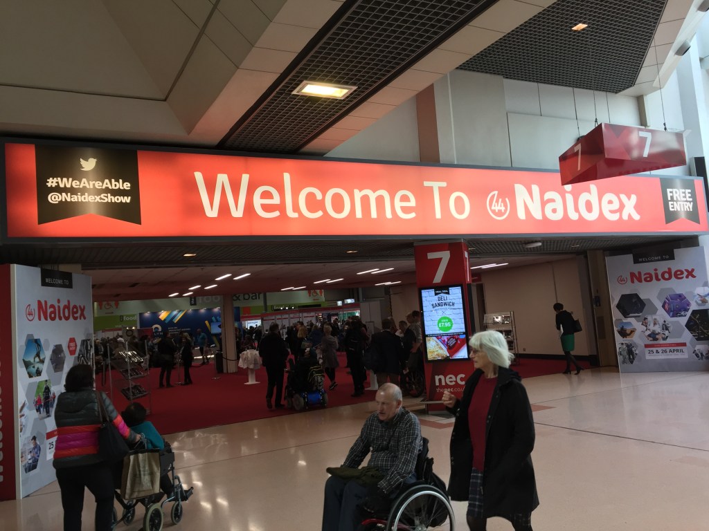 Entrance to Naidex show. Above a wide entrance to the hall is a long red sign, on which is large white text saying Welcome To Naidex. White text on a black panel to the left shows their Twitter name, Naidex Show, and the hashtag We Are Able. White text on a black panel on the right says Free Entry.