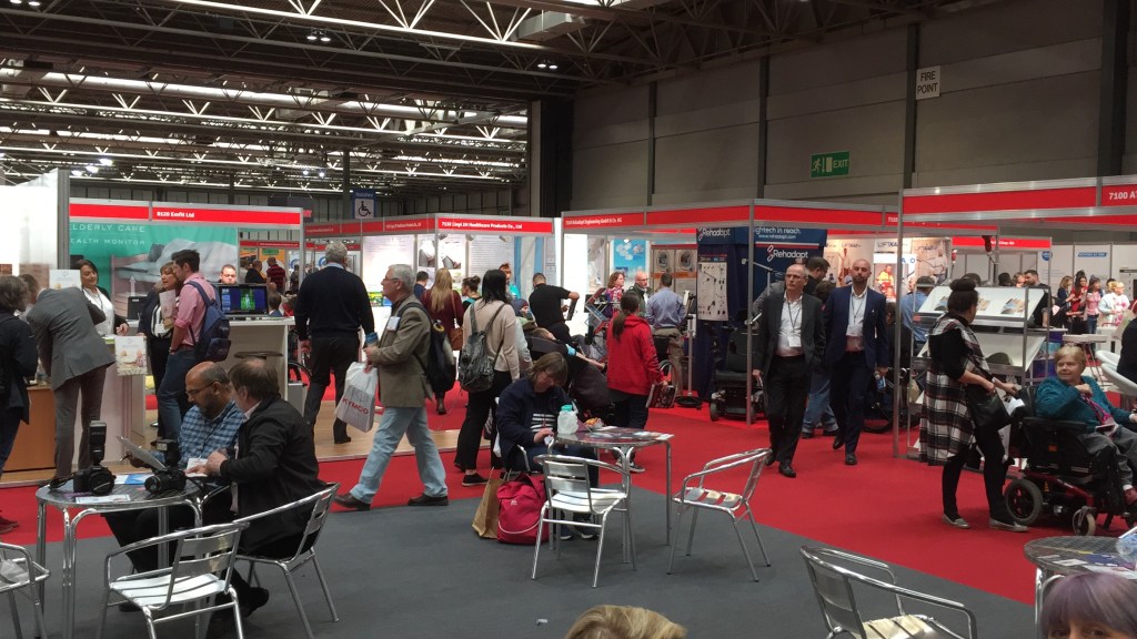 Lots of people walking around the various exhibitor cubicles in the Naidex show. Each cubicle has a narrow red banner across the top with the stand number and exhibitor name in white text.