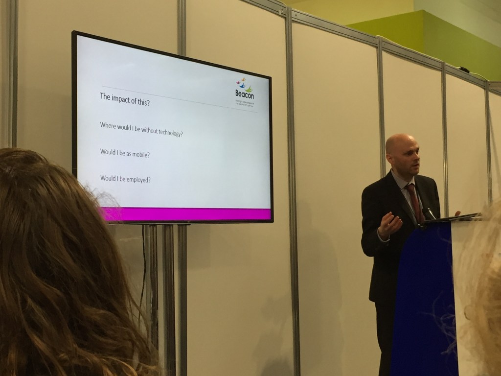 Matt giving his talk standing at a lectern. On a screen next to him, a Powerpoint slide reads