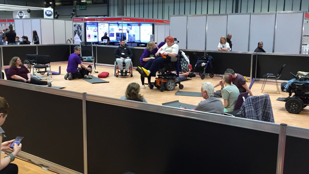People on mats and in wheelchairs doing yoga at Naidex while others watch