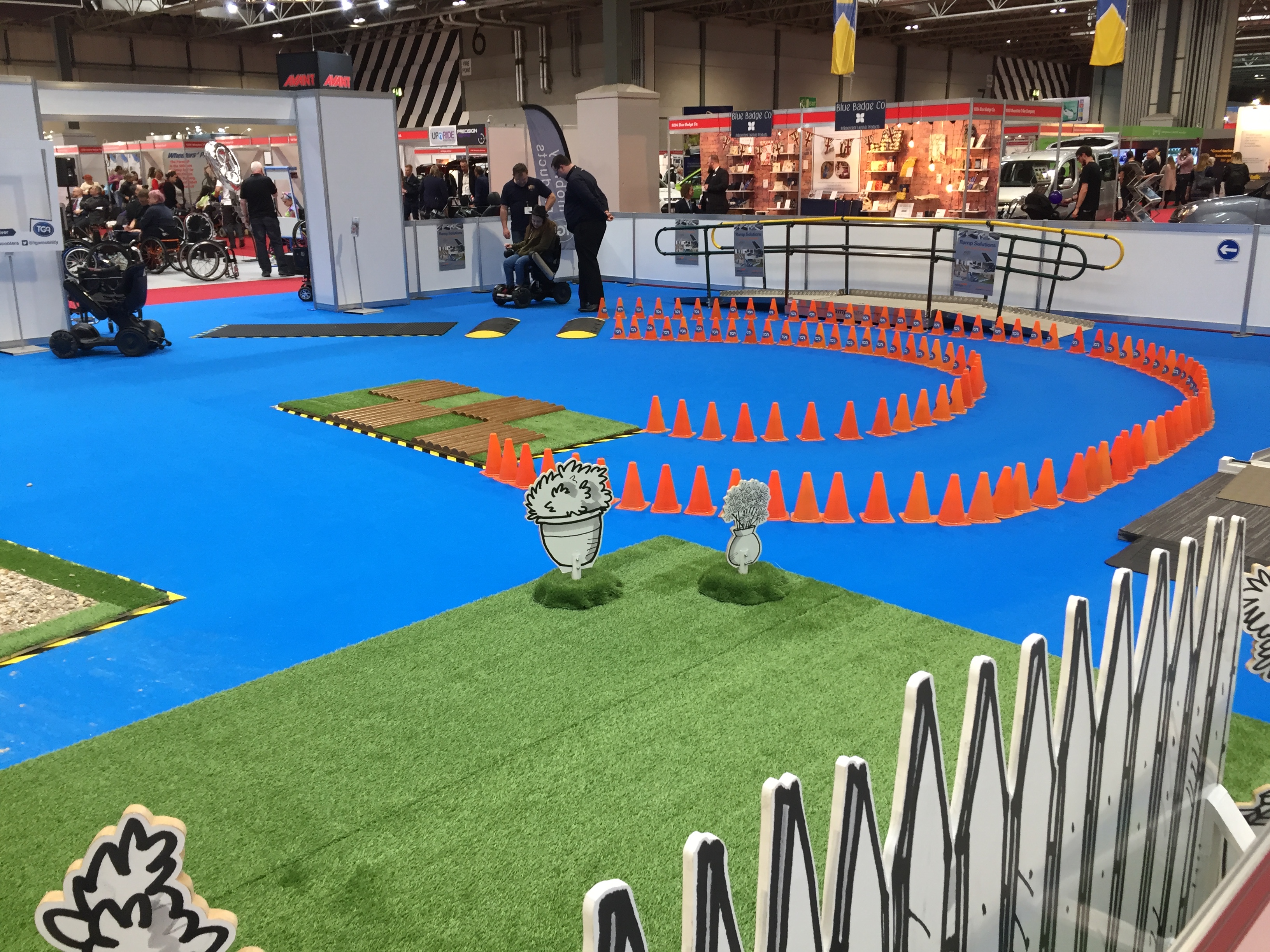Right side of the Mobility Test Track at Naidex. On a large blue floor are 2 small speed bumps leading to a curved path created by cones, leading a section of uneven paving. There is also a large patch of fake grass to try driving on.