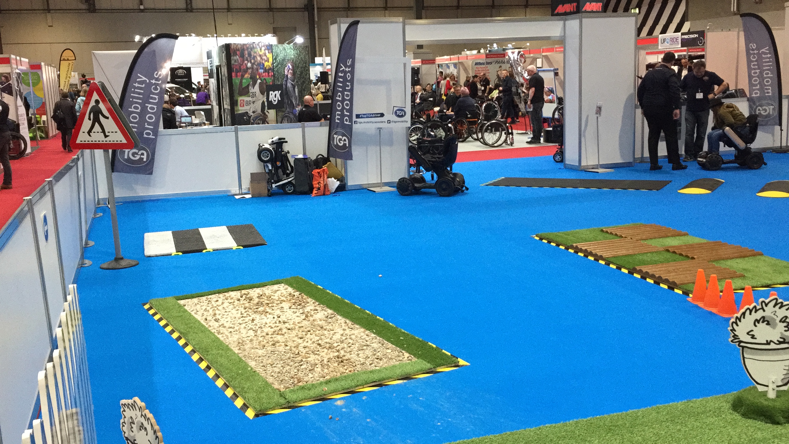 Left side of the Mobility Test Track at Naidex. On a large blue floor are obstacles including a small mockup of a zebra crossing, a section of gravel, and a section of uneven paving.
