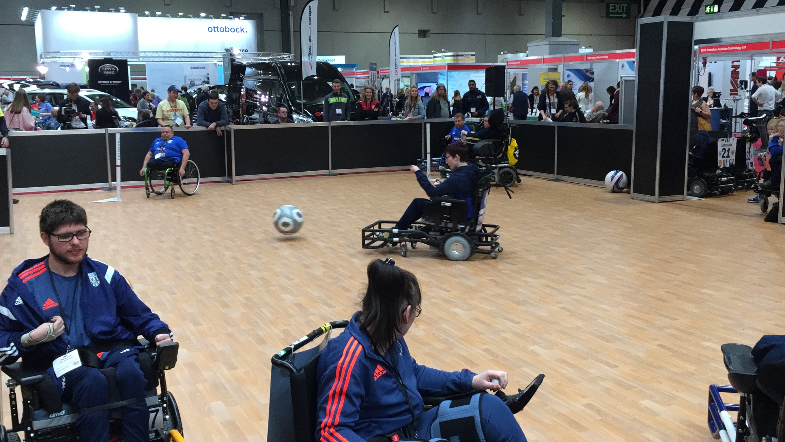 Powerchair Football at Naidex, with the ball travelling towards the goal, having been kicked by one powerchair user.