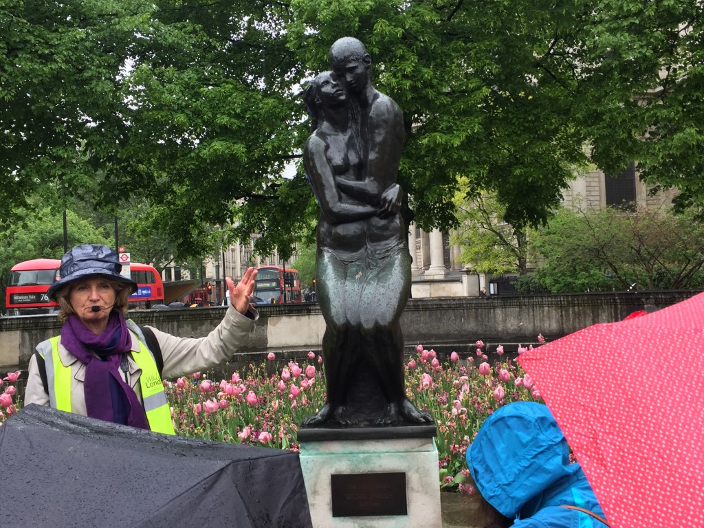 Tour guide gesturing to a tall statue of a girl and boy embracing as they lovingly look into each other's eyes.