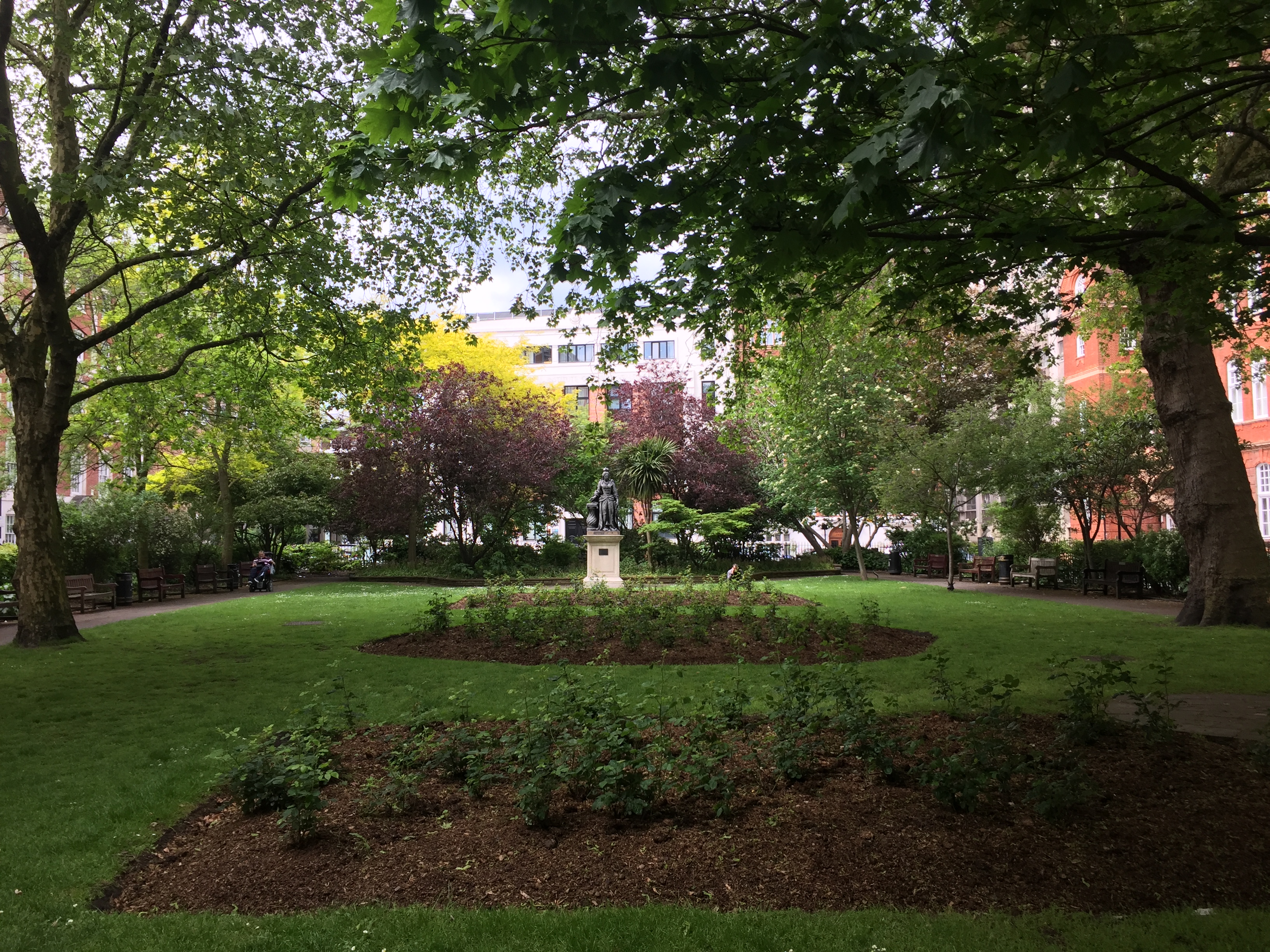 A section of Queen Square Garden, with a round flowerbed in the middle of the grass, and behind it a statue of Queen Charlotte