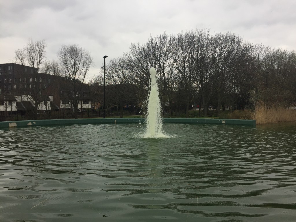 A tall fountain in a large round pool in Mile End Park.