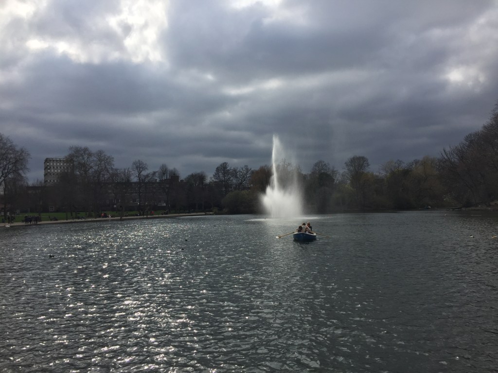 A large fountain spraying high in the West Lake in Victoria Park, while a family of 2 adults and 2 children are in a rowing boat nearby.