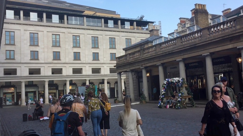View of Covent Garden Square, looking towards a tall white building on the corner ahead. The big market building on the right, which forms the centre of the square, is lined with pillars, and the space behind them is dark.