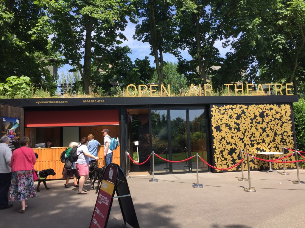 Exterior of the Open Air Theatre building, with the booking office desk on the left, the entrance door in the centre, and a wall covered in gold leaf detailing on the right. On top of the single-floor building are letters that spell out Open Air Theatre.