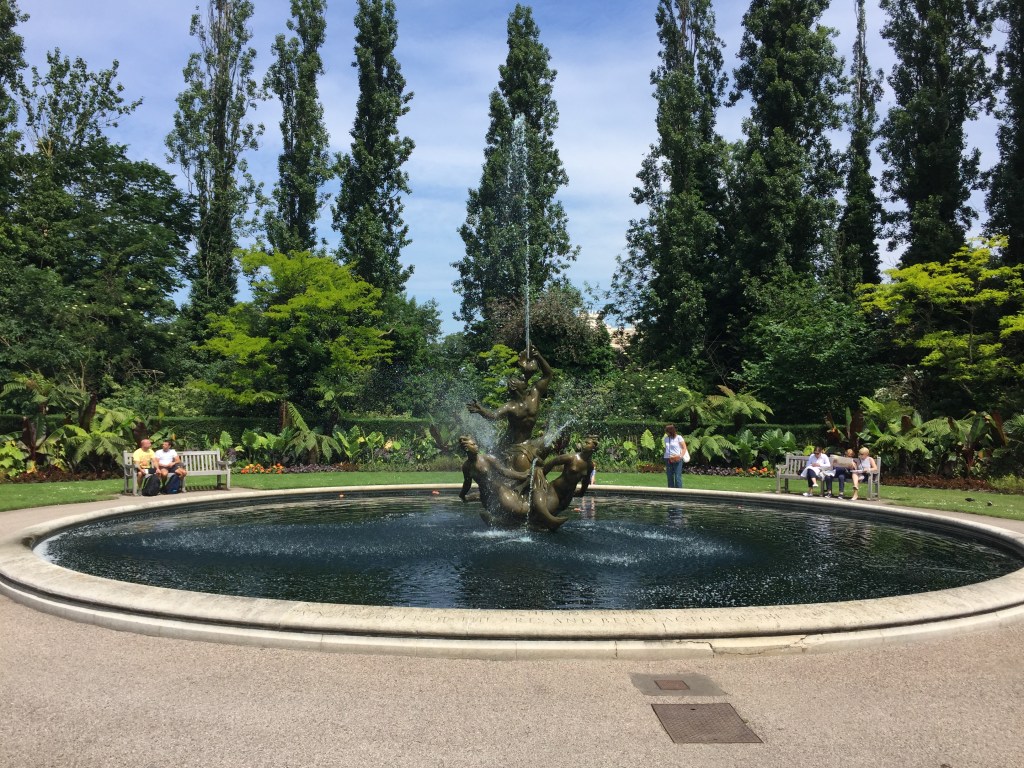 Large circular pool with a fountain statue in the middle. The statue is made of 3 figures - one male figure in the centre looking up and spraying a jet of water high into the air, and then 2 female figures either side of him, spraying water out in various directions around them.
