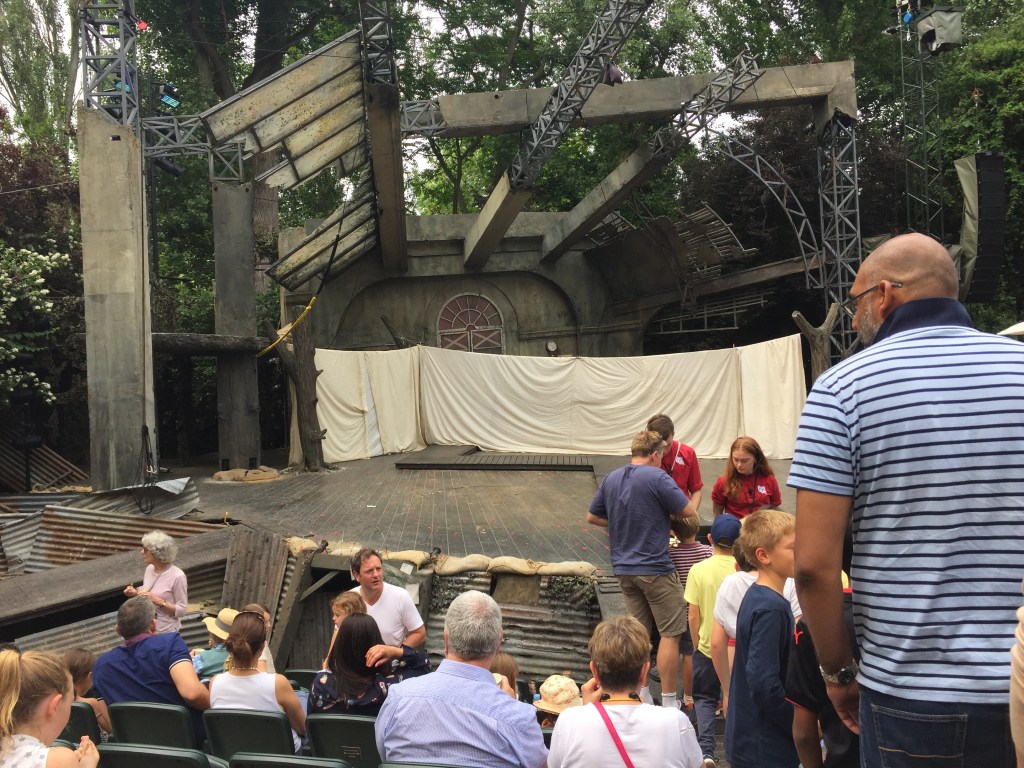 A view of the theatre stage, which now has a long wall made of sheeting across the back of the stage, made by sheets draped over upturned beds. Next to where we're sitting, audience members are queuing down the aisle for small tubs of ice cream being sold by 2 members of staff at the bottom of the steps by the stage.