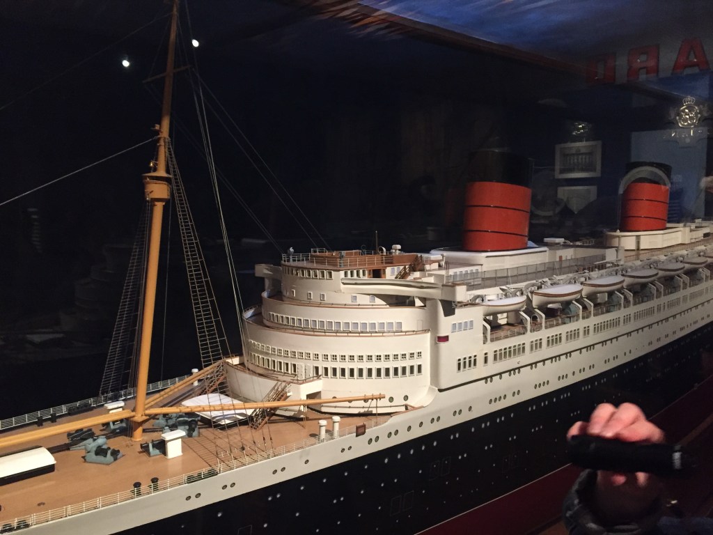 A very long and highly detailed model of the Queen Elizabeth Cruise Liner. The ship has a black base, with white buildings on the top, and details including the large mast down to the small lifeboats, windows and railings are all reproduced in the model.