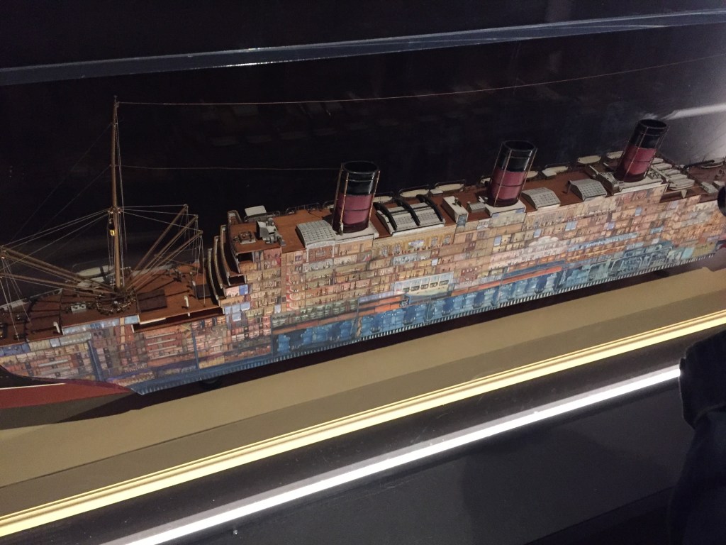 Large cross section model of an ocean liner. The side of the boat has small images of all the rooms on that side of the vessel, including items of furniture and people in the rooms.