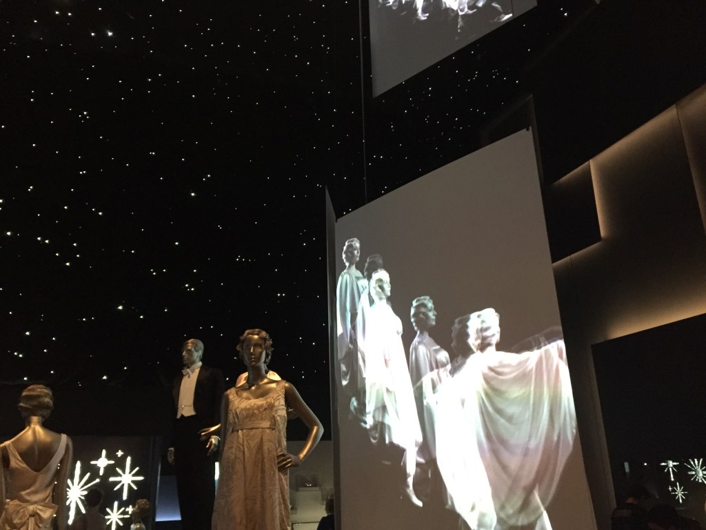 Below a black ceiling covered in stars to look like the night sky, a large screen shows black and white footage of ladies in long dresses descending a staircase. In front of the screen are mannequin models of a man in a smart suit and a woman in an elegant dress.