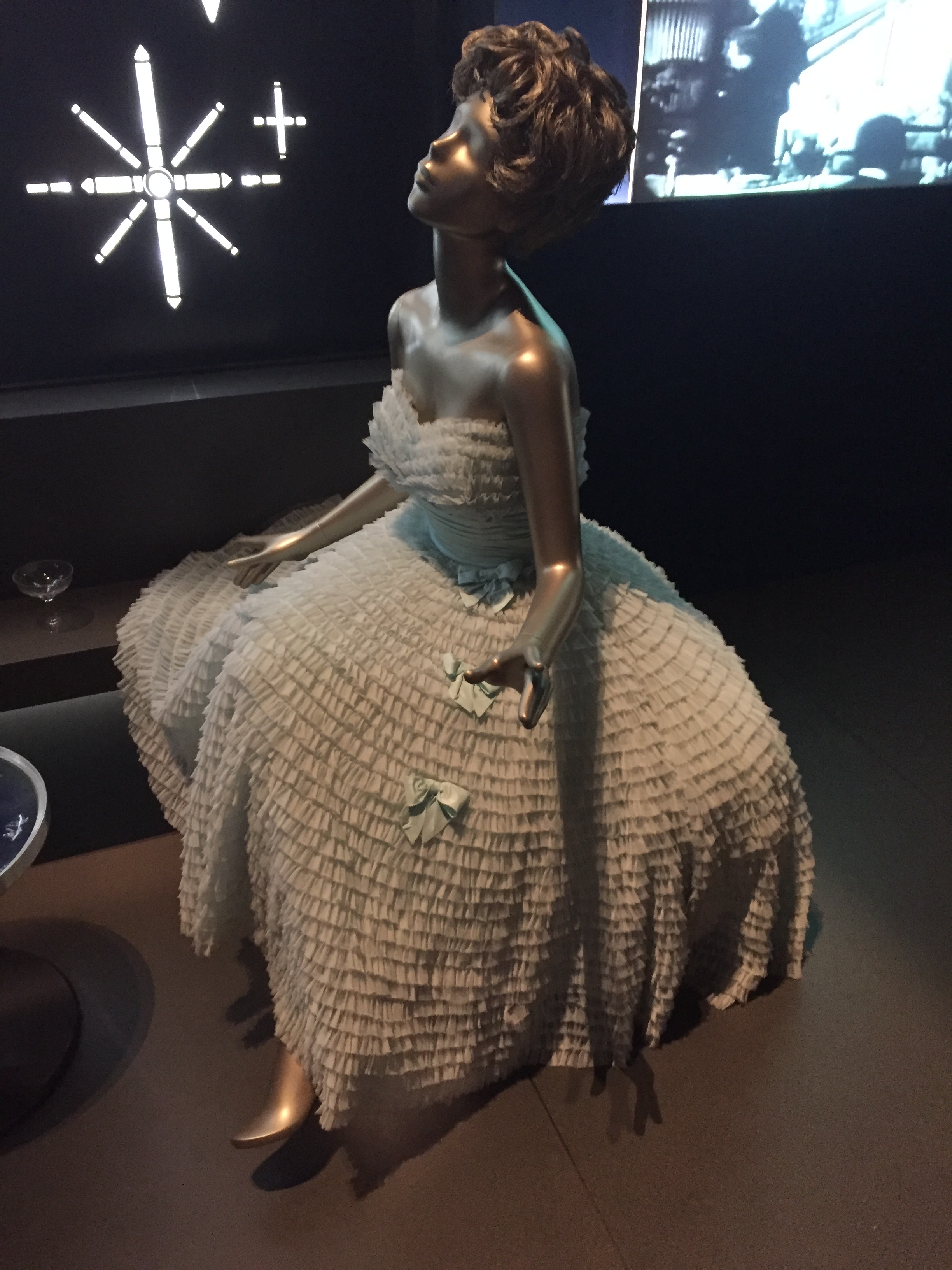 Mannequin wearing a white dress that spreads out widely at the bottom, covered in many layers of ruffles and adorned with a few bows.