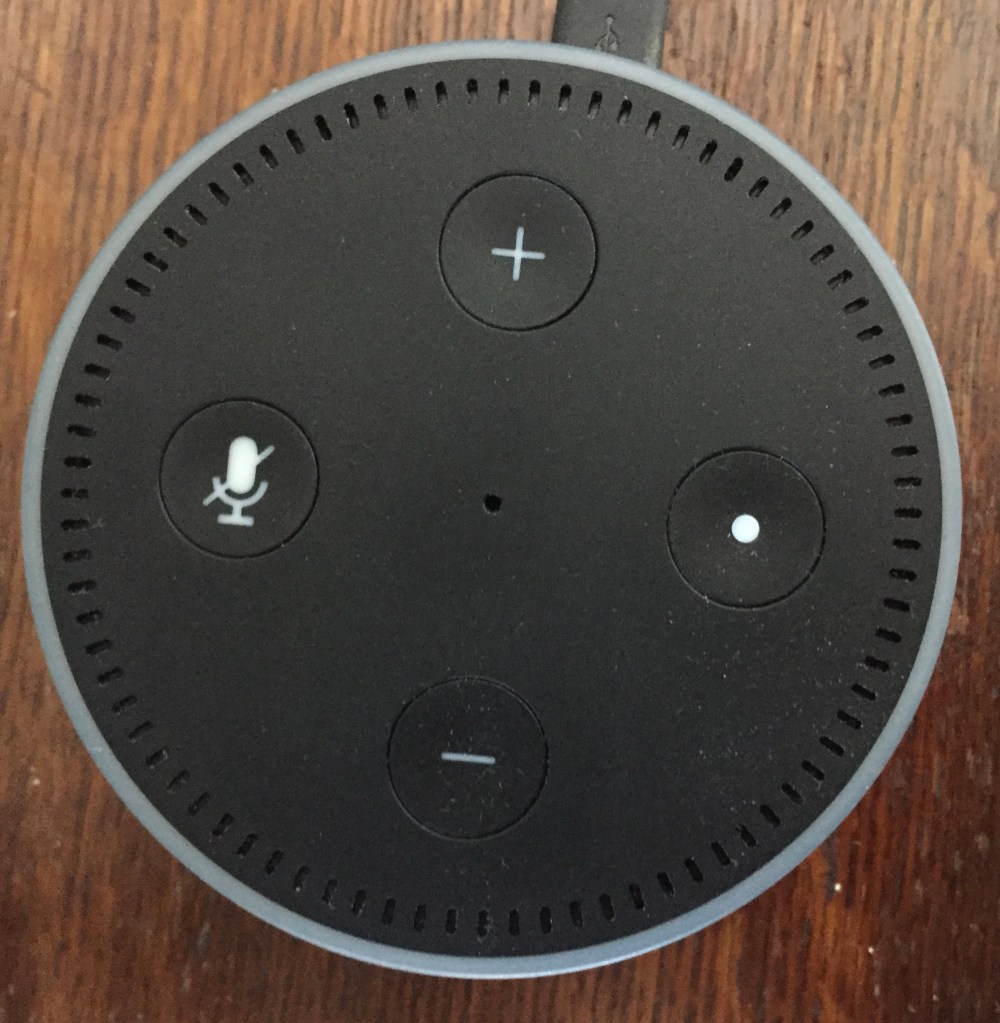 Amazon Echo Dot - Small round black machine with a small speaker grill and light encircling the outer edge on top. 4 buttons are on the front - plus and minus volume buttons at the top and bottom, a mute button on the left, and a control button with a dot on it on the right.
